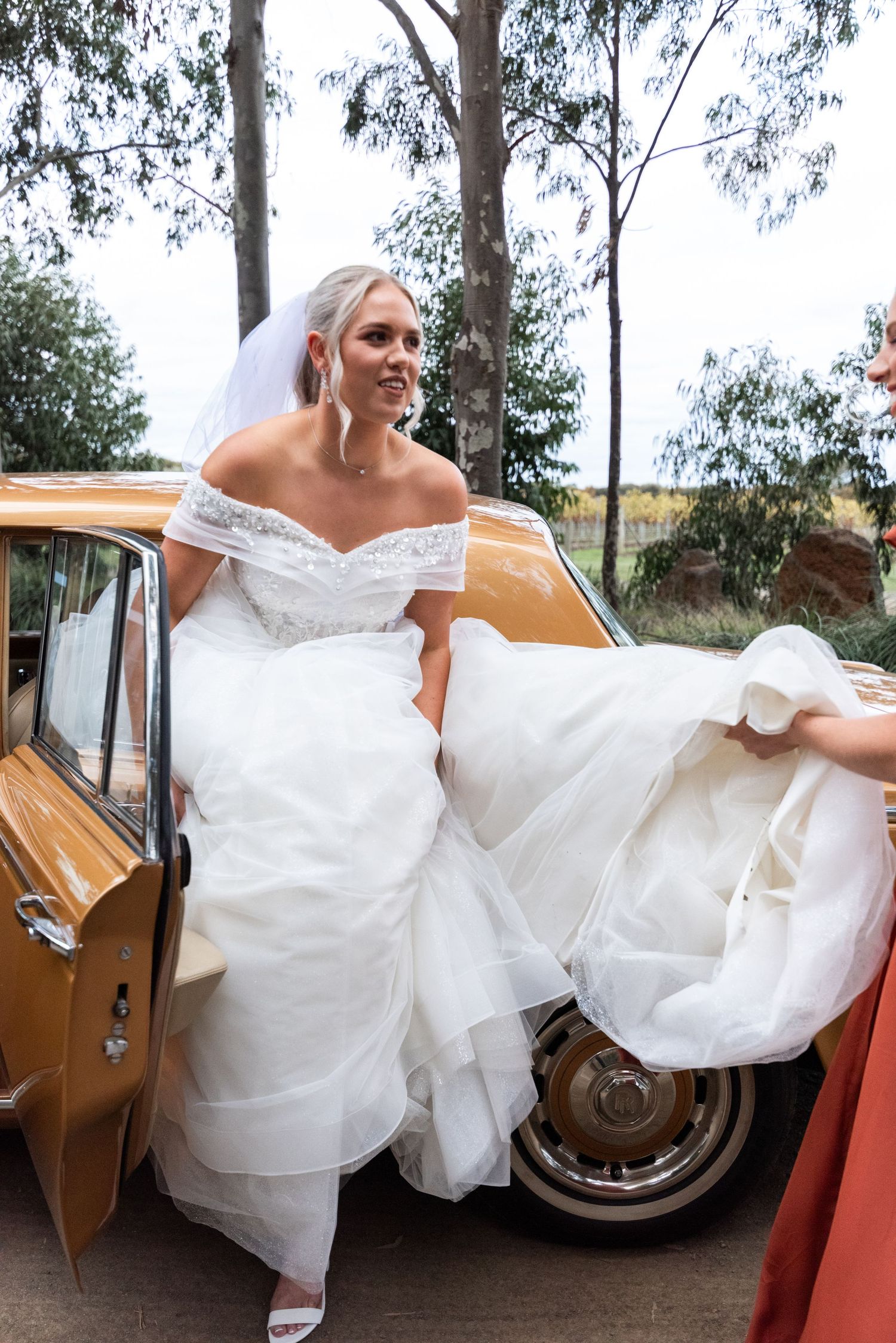 Bride in off-shoulder wedding dress poses elegantly next to vintage orange car.