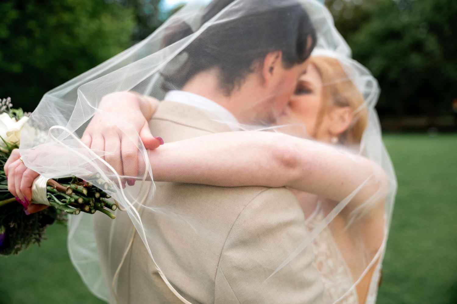 Artistic wedding portrait with flowing veil against green garden backdrop.