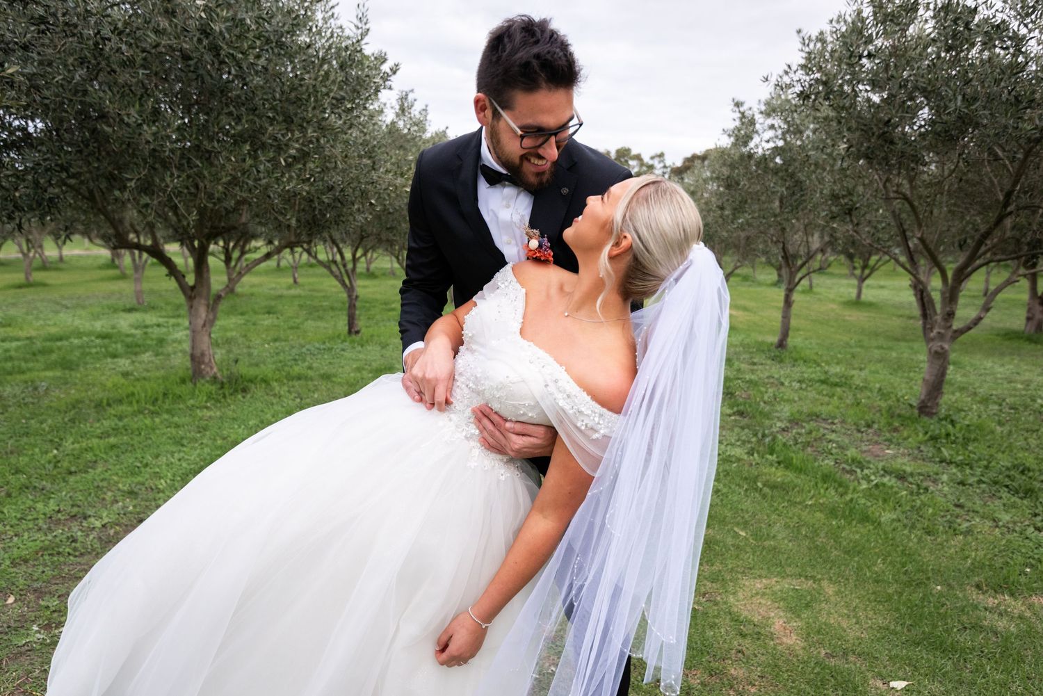 A romantic wedding photo of a couple embracing in an olive grove, with the bride wearing a flowing white gown and veil.