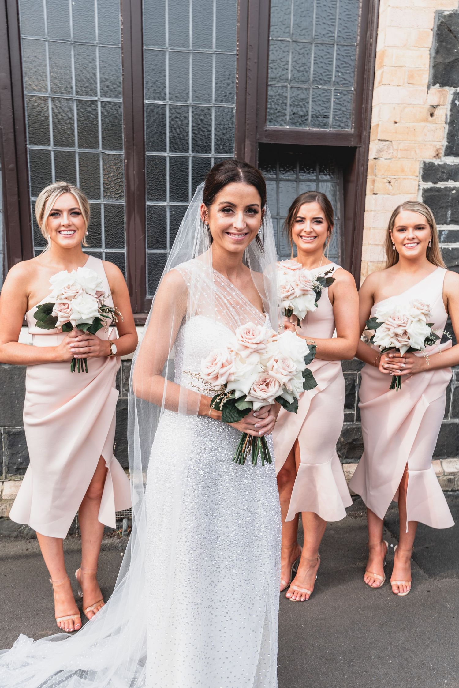A wedding party poses in blush pink bridesmaid dresses and a white wedding gown outside a brick building.