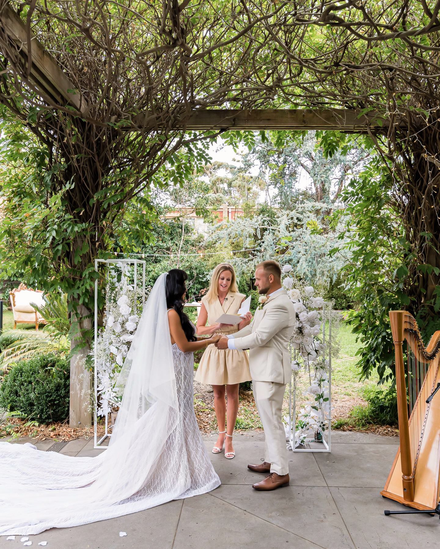A wedding ceremony takes place under a lush green garden archway with musicians and guests gathered around.