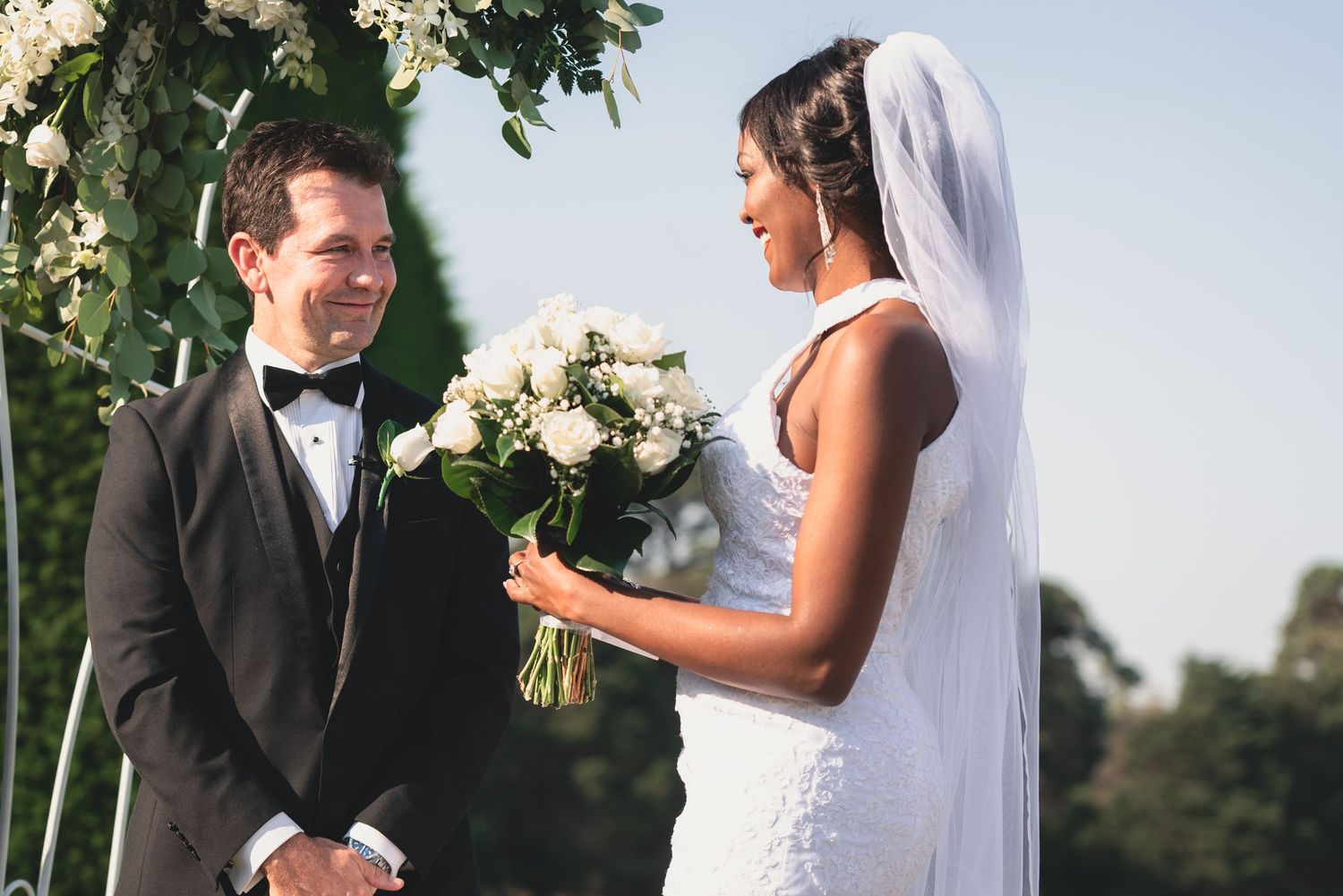 A bride and groom share an intimate moment during their outdoor wedding ceremony among white floral decorations.