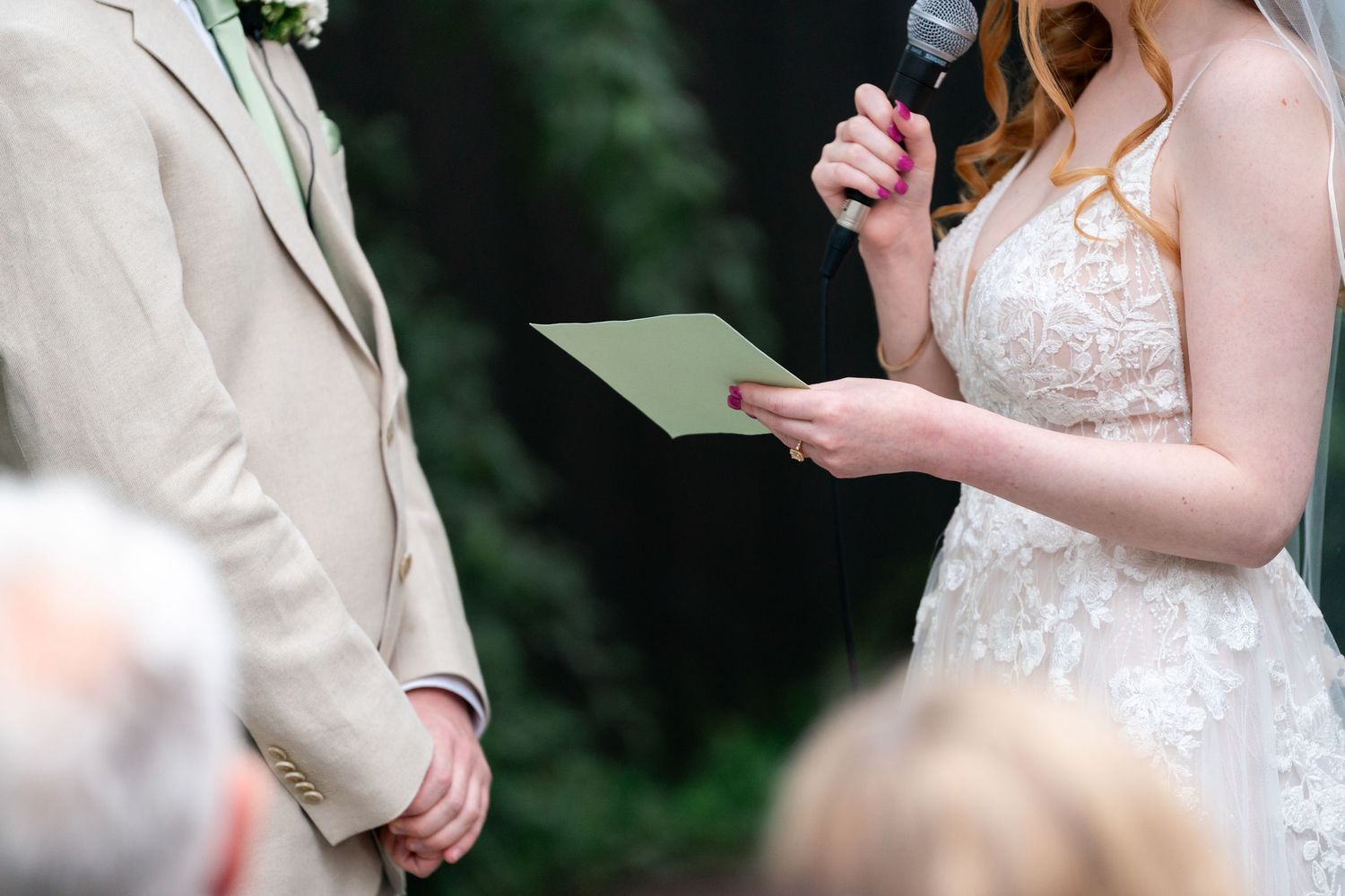 A bride in a white lace dress reads wedding vows from a paper while holding a microphone during an outdoor ceremony.