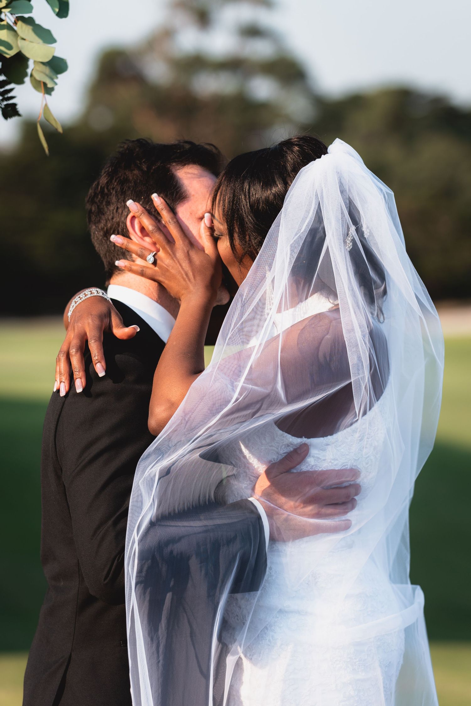 A romantic wedding couple shares an intimate kiss outdoors under a flowing white veil in natural lighting.