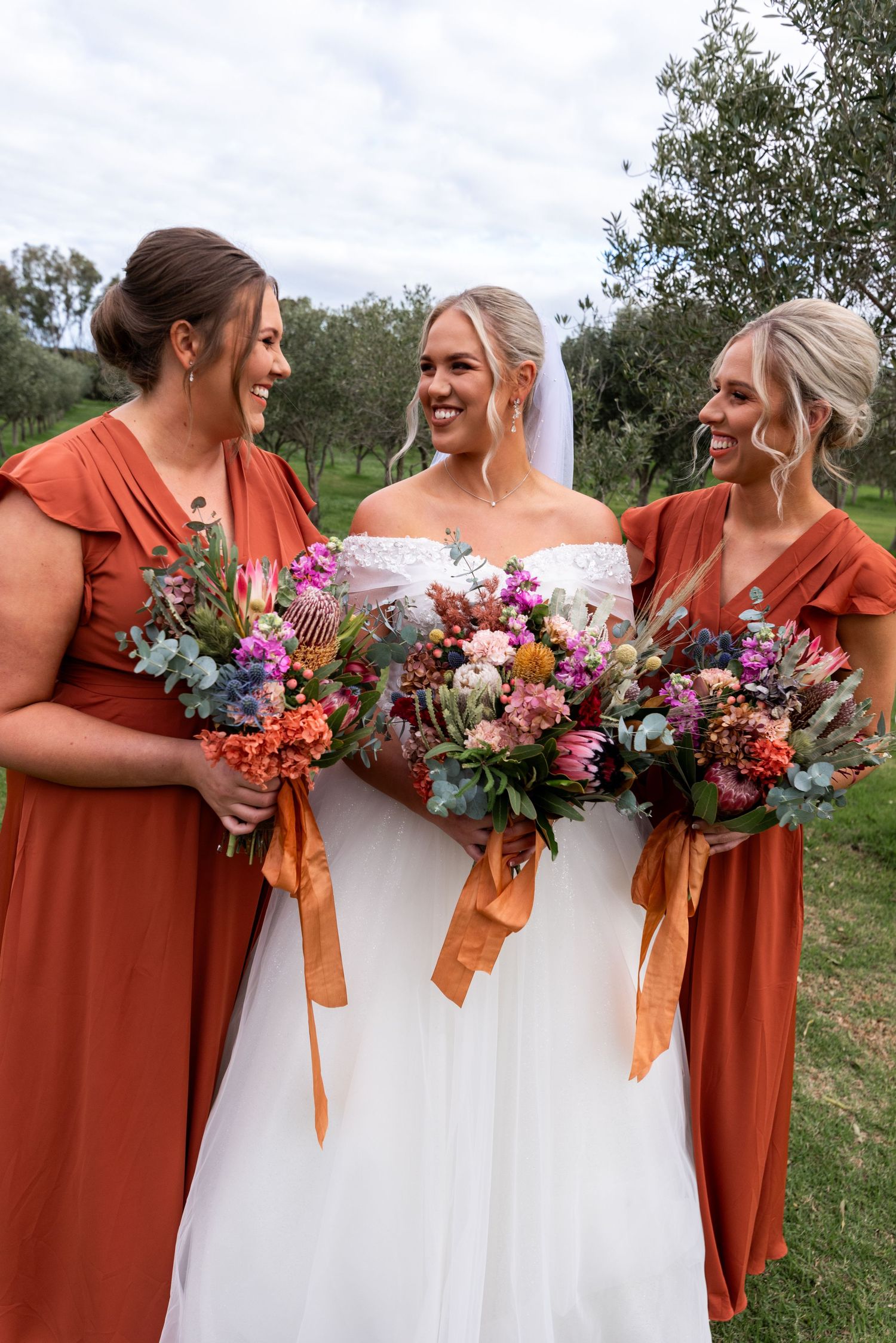 Bridesmaids in rust-colored dresses hold vibrant bouquets with orange ribbons next to bride in white gown.