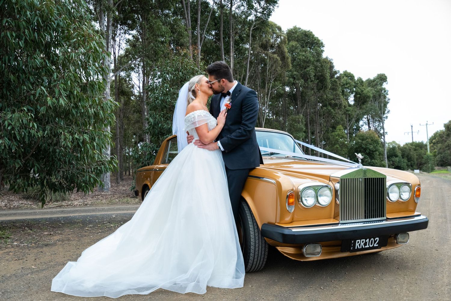 A wedding couple poses beside a vintage gold Rolls Royce car in a wooded setting.