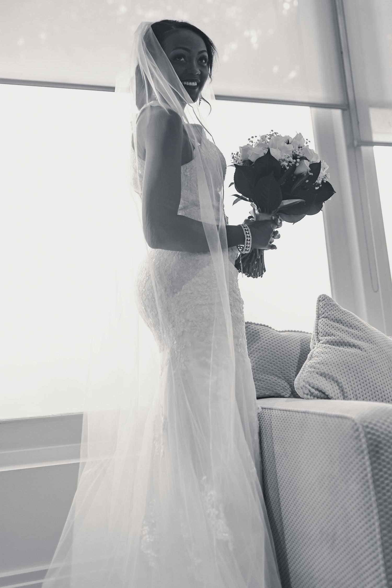 A bride in a fitted lace wedding gown holds a white floral bouquet while standing near a window in black and white.