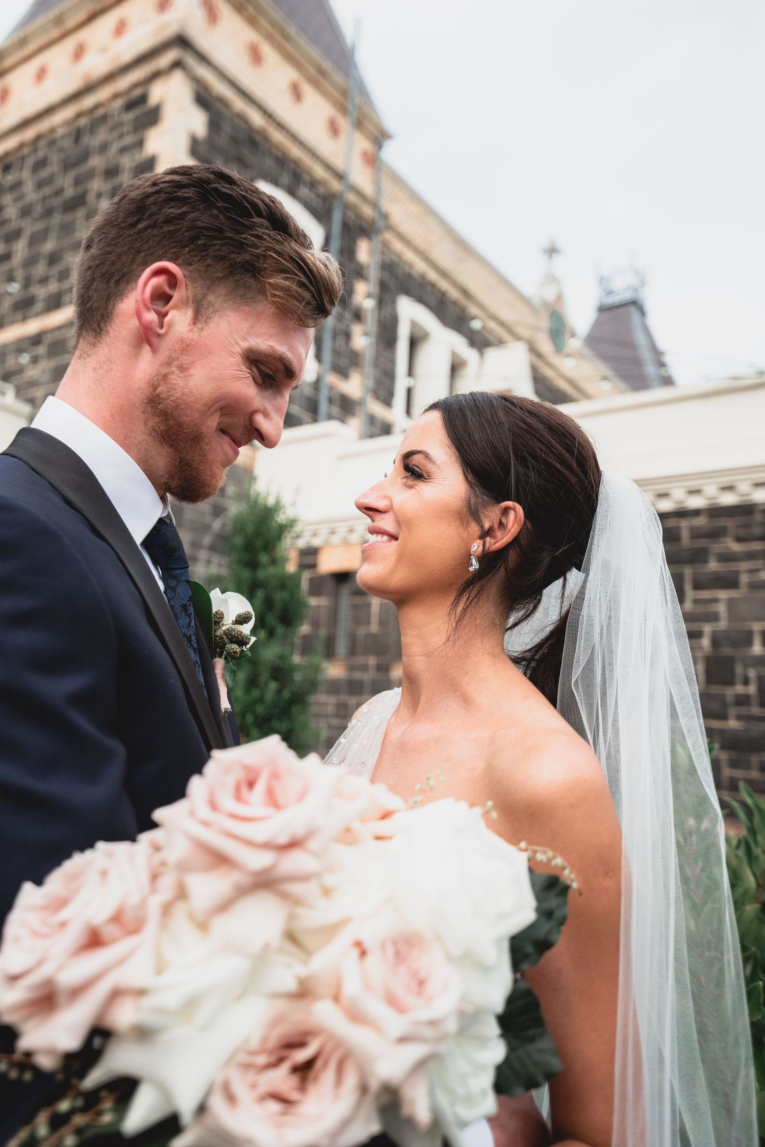 A happy couple shares intimate moments in their wedding attire against a stone building backdrop.