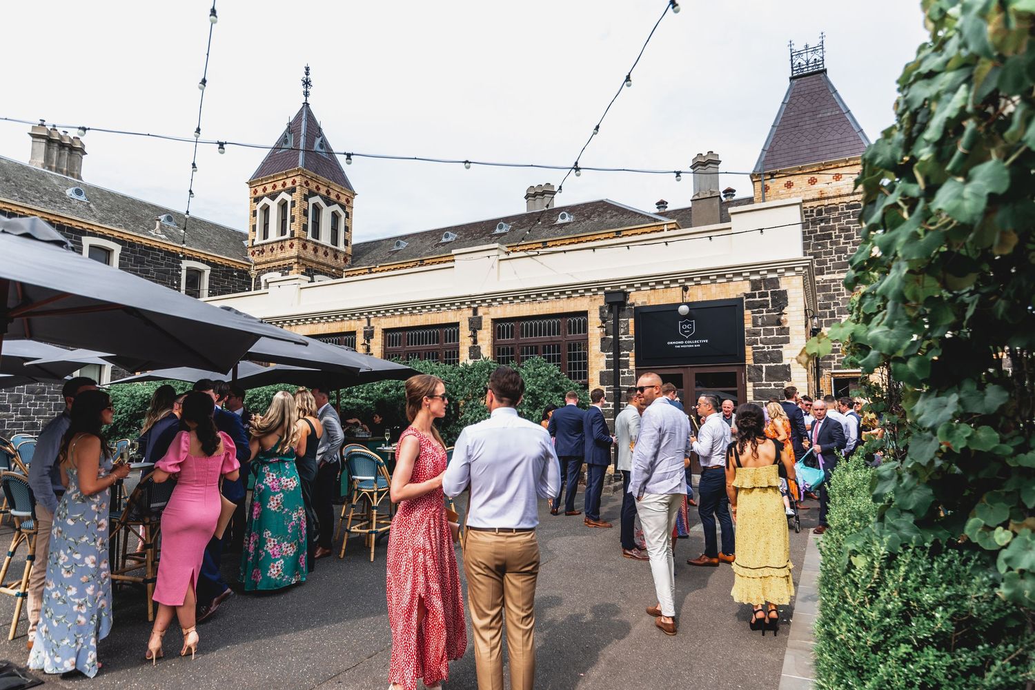 Wedding guests in colorful summer attire gather at an outdoor venue with stone architecture and greenery.