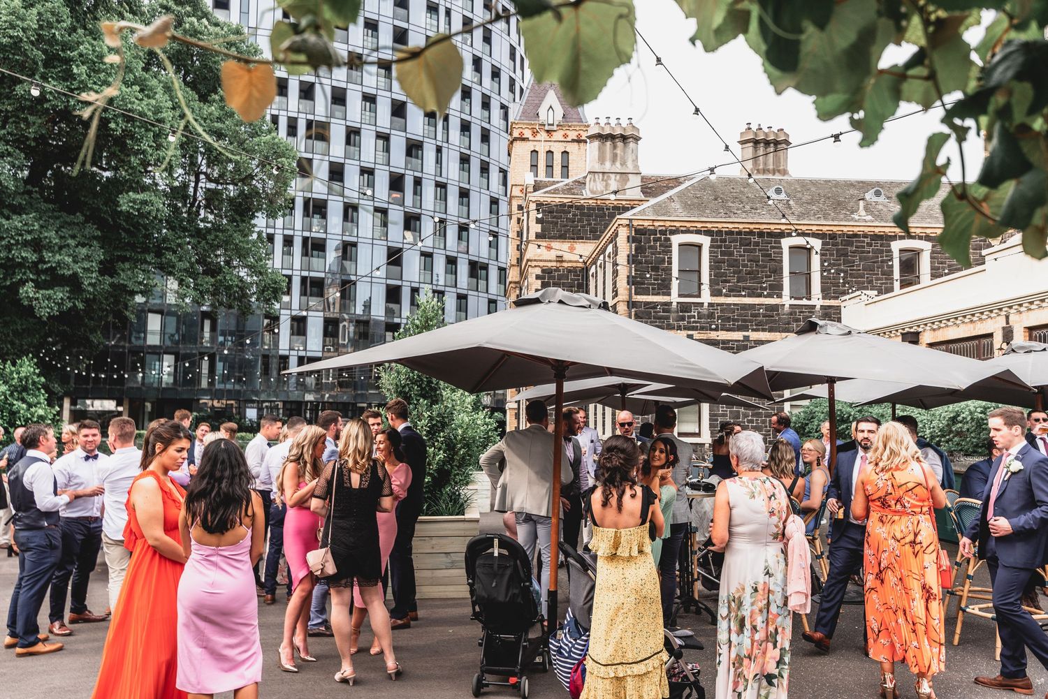 A crowd of people in summer dresses gather at an outdoor event with modern architecture and stone buildings in the background.