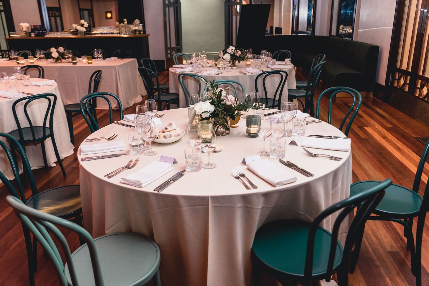 Elegant round dining tables with white tablecloths and green chairs set for a formal dinner service in a restaurant.