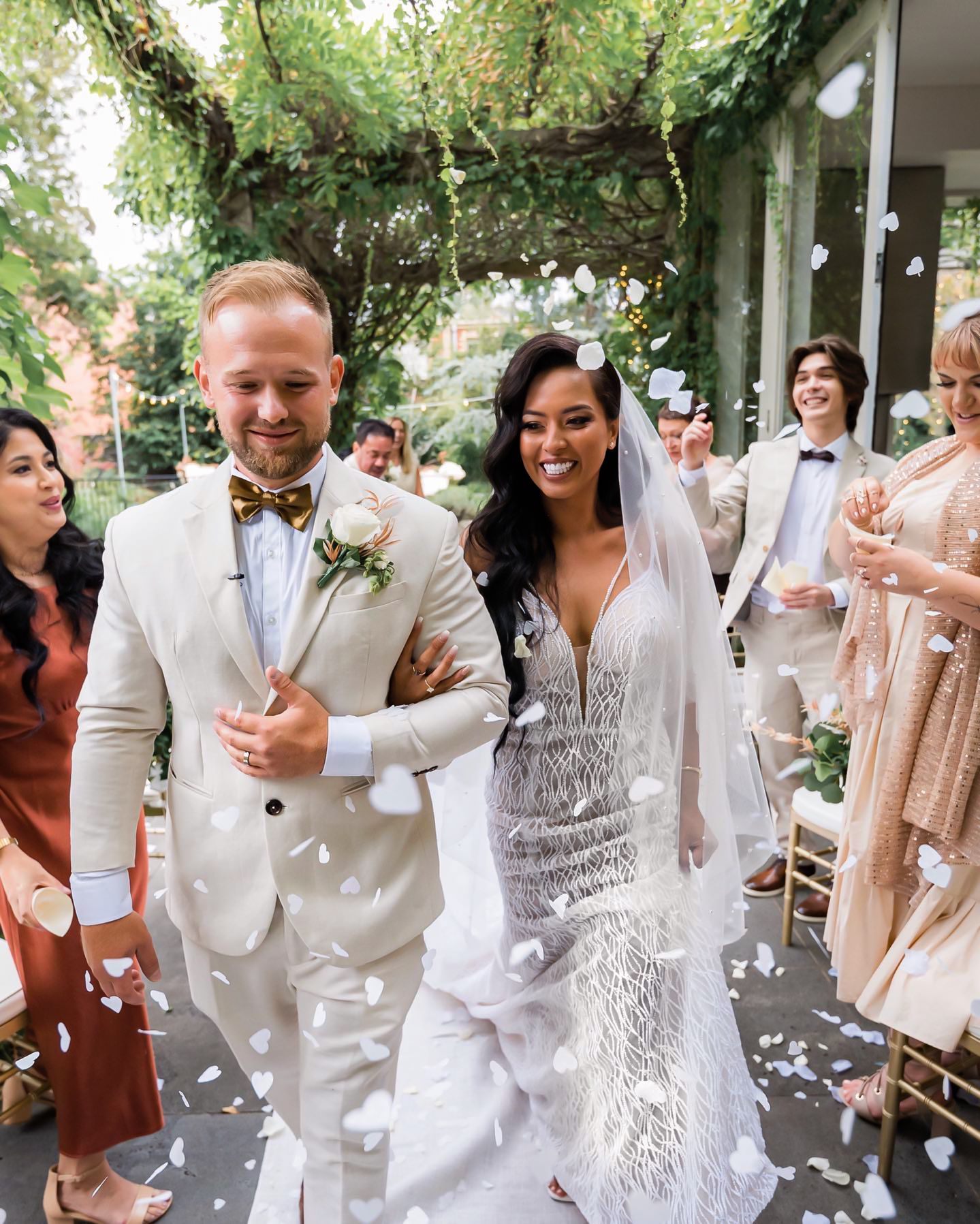 Wedding party celebrates outdoors with joyful dancing and festivities under green trees and string lights.