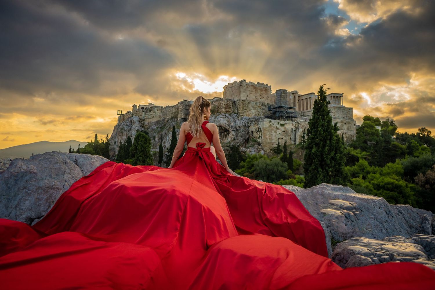 Billowing red ball gown creates stunning silhouette against Athens Acropolis at sunset.
