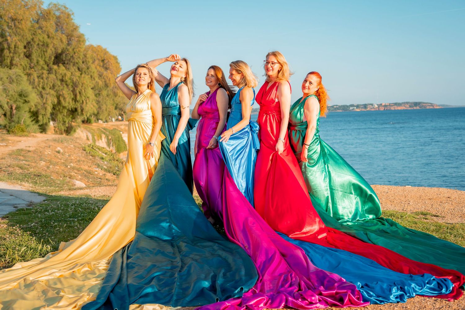 Group wearing vibrant flowing dresses in yellow, blue, purple, red and green poses along a scenic beach at sunset.
