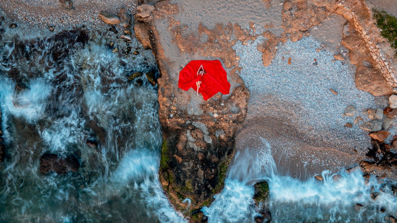 drone view of red flying dress on coastal rocks with turquoise waters swirling below.