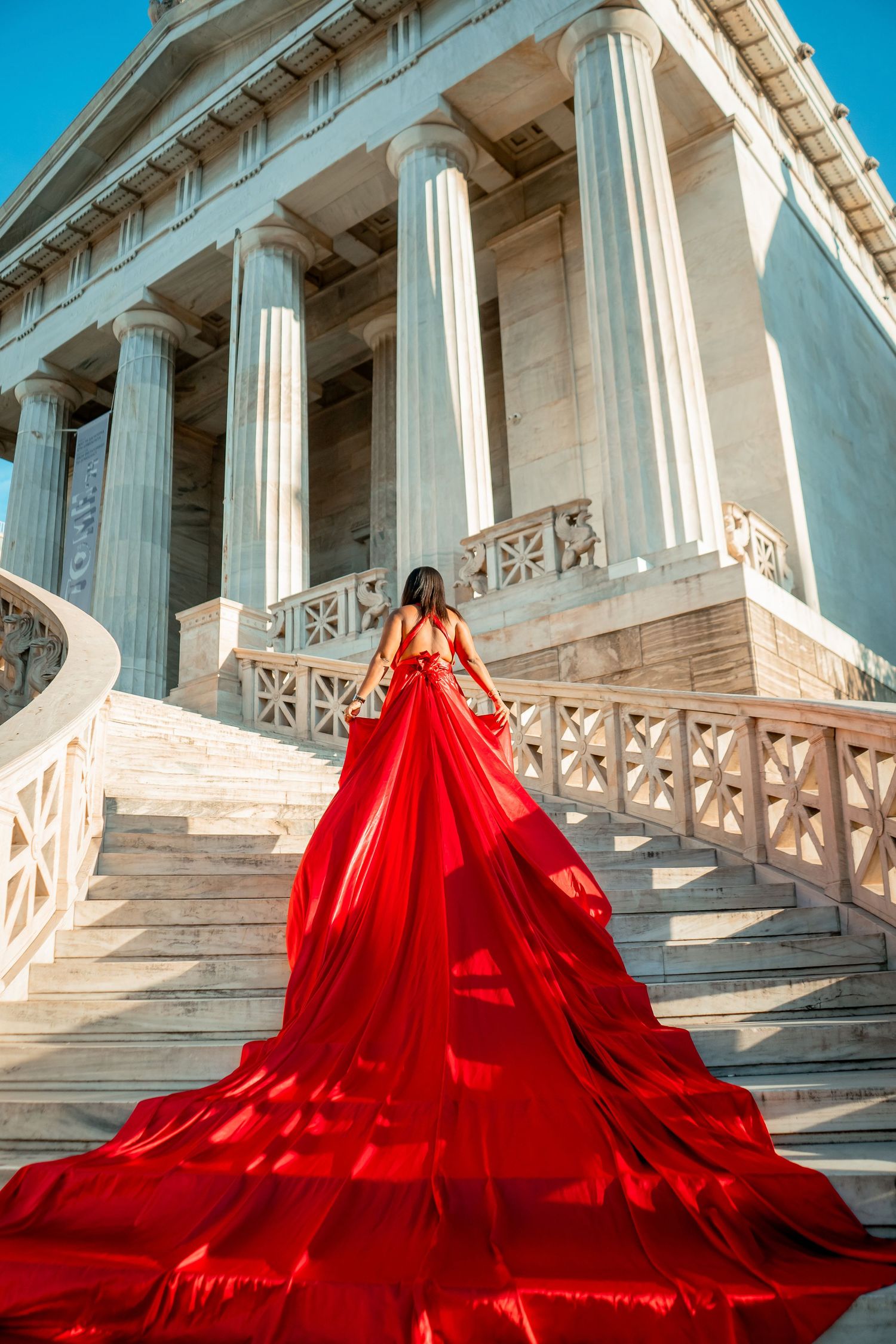 Dramatic red ball gown with long train on white marble steps of classical building with columns.