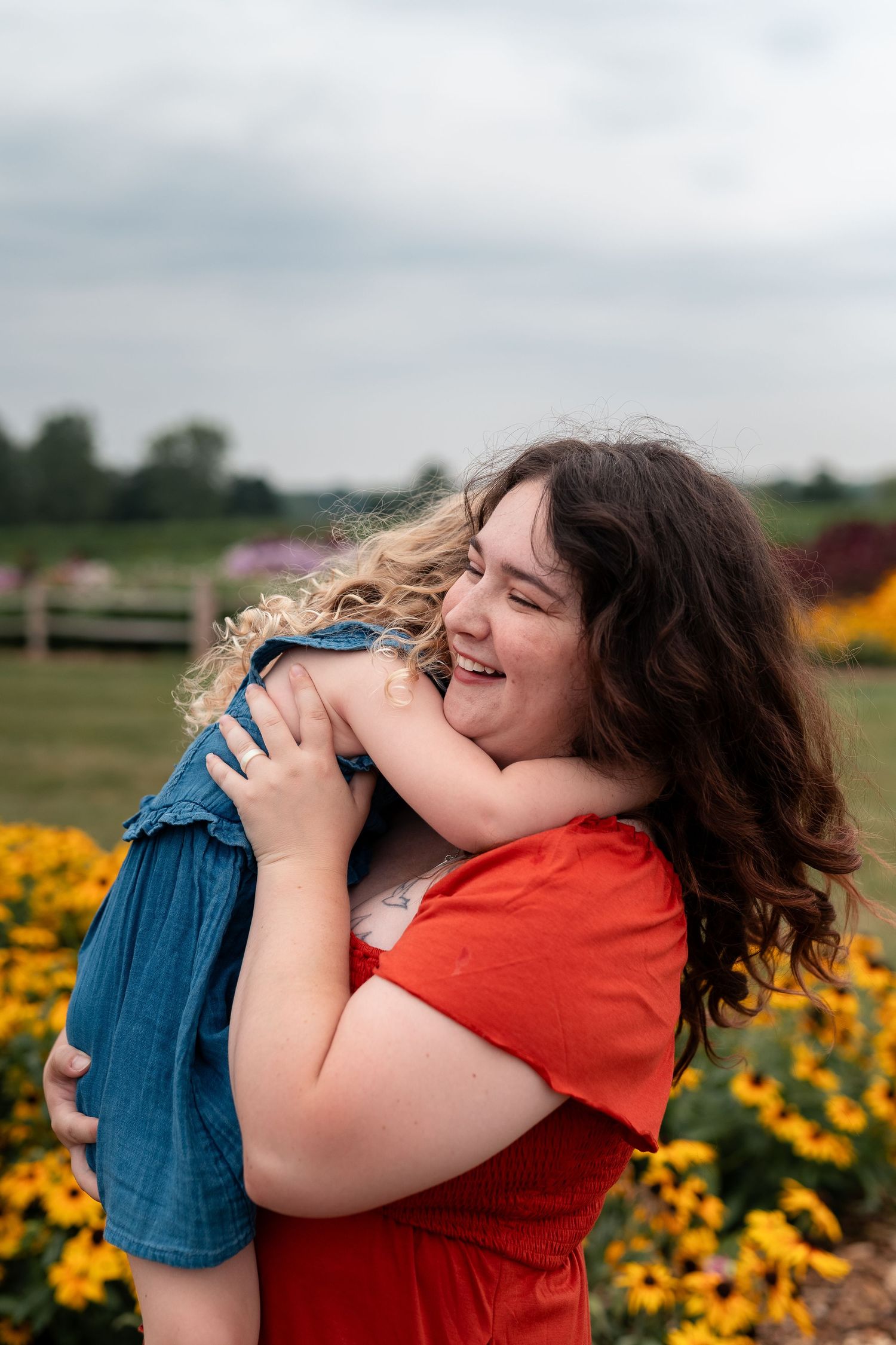 A smiling woman in a red dress hugs a young child in a denim outfit among yellow flowers outdoors.
