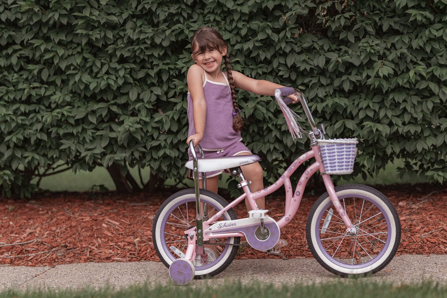 Child riding a white bicycle with training wheels along a path bordered by green foliage.