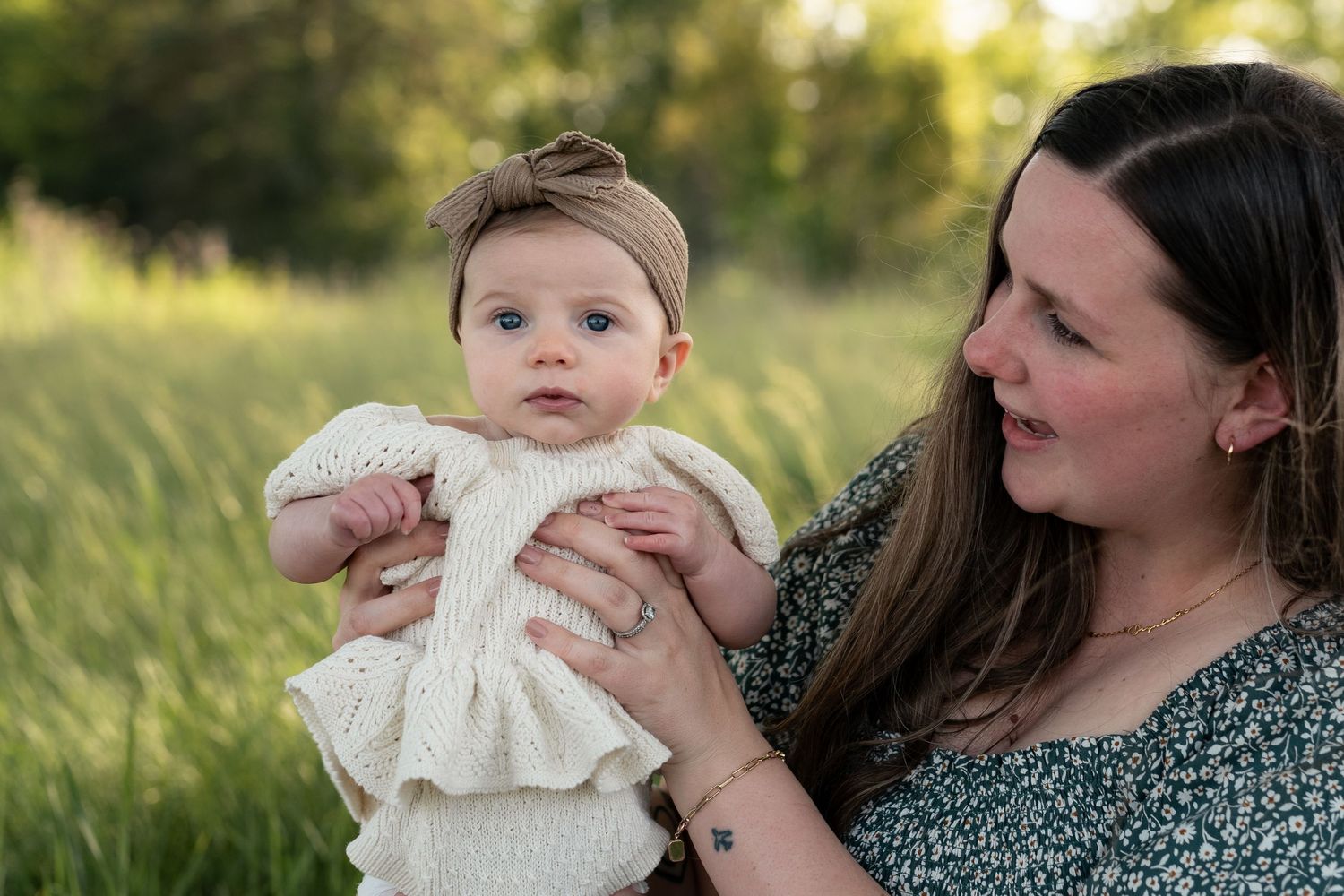 A mother holds her baby girl in a white knit dress outdoors in a green field during golden hour.