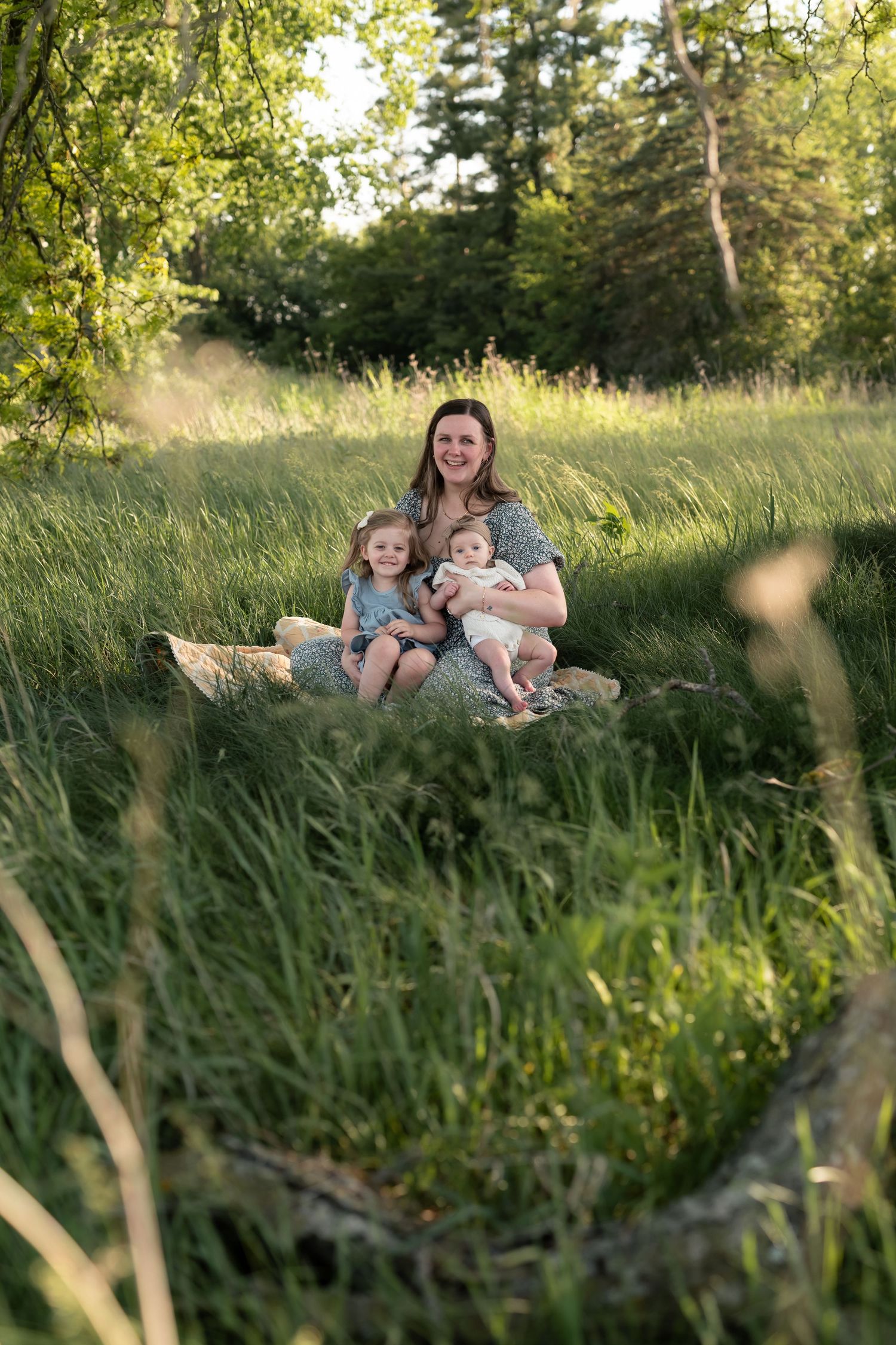 A smiling mother sits in a lush green meadow holding a baby and toddler on a sunny day.