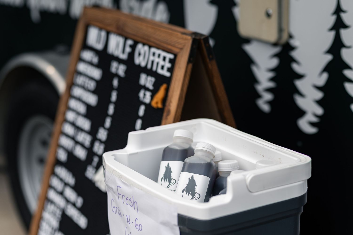 Wild Wolf Coffee cold brew bottles in a cooler beside a chalkboard menu sign at an outdoor coffee stand.