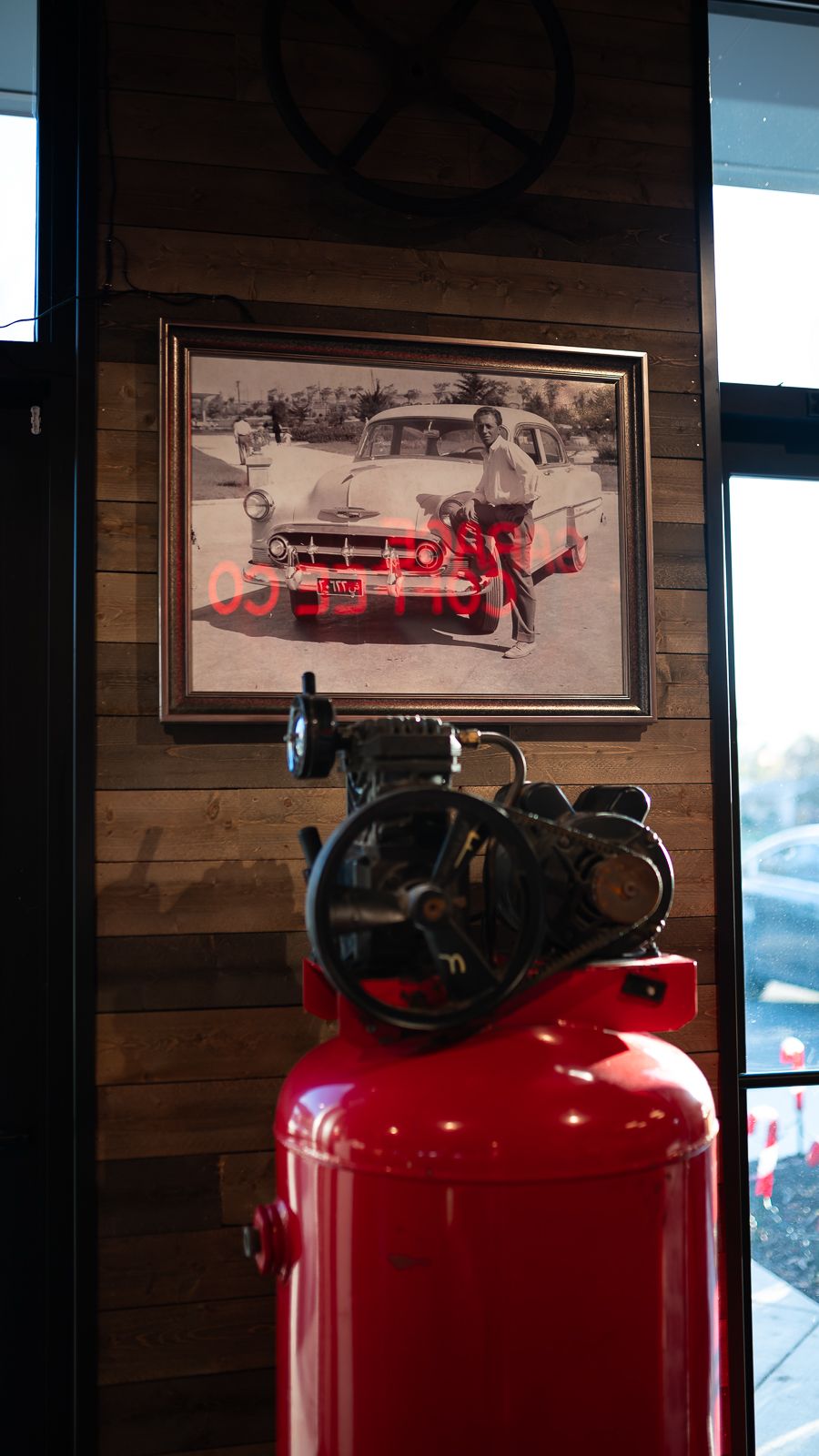 Red vintage air compressors displayed against a wooden wall with black and white automotive photographs above.