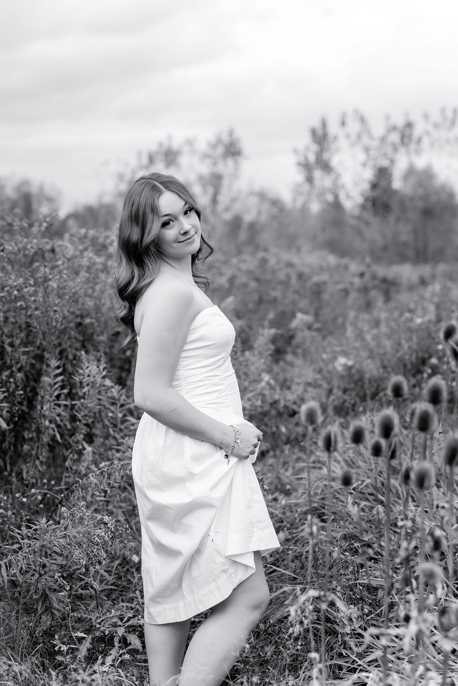 Black and white portrait of young woman in white dress standing among tall wildflowers in field.