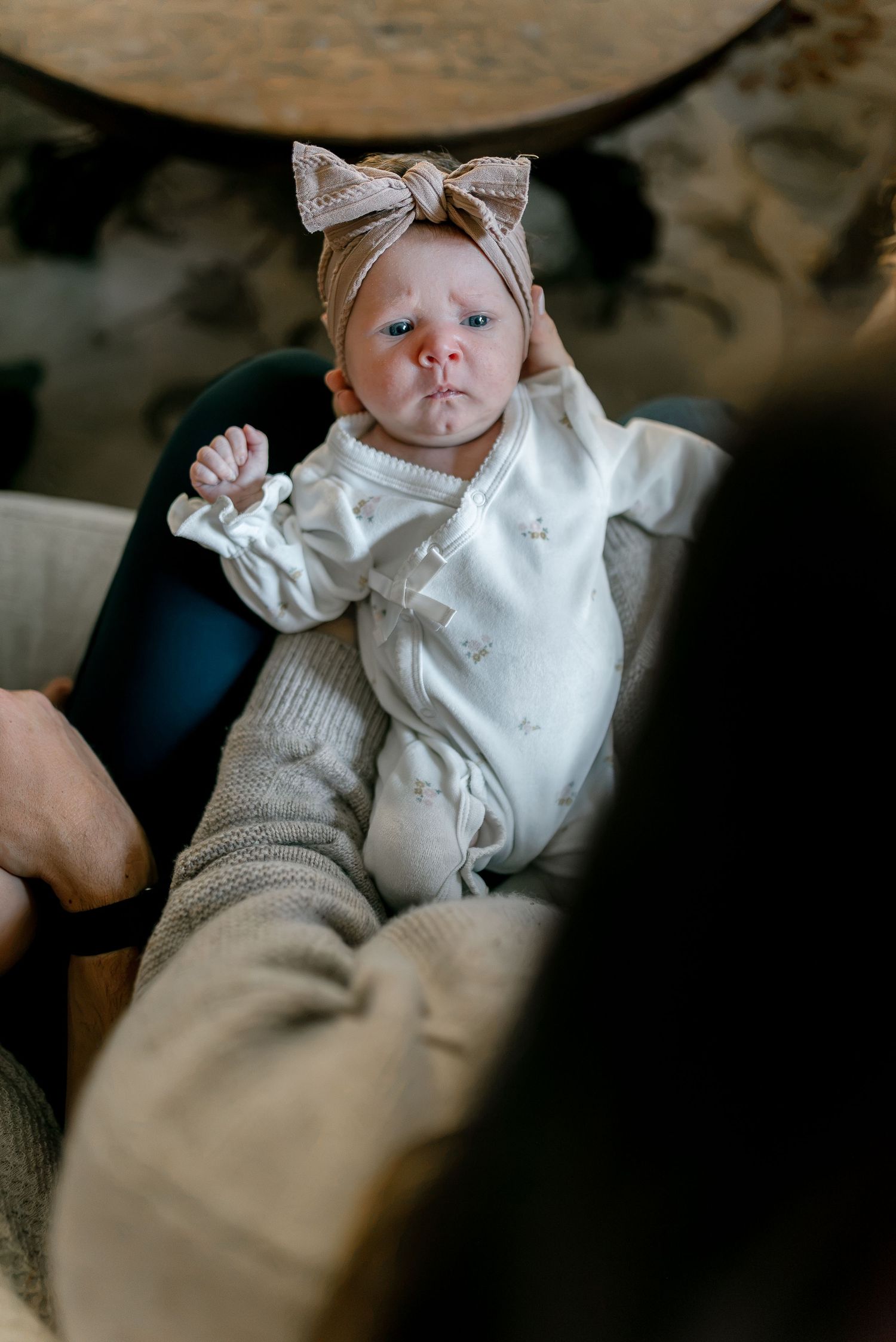 Newborn baby girl in white outfit and beige bow headband lying in adult's arms looking at camera.