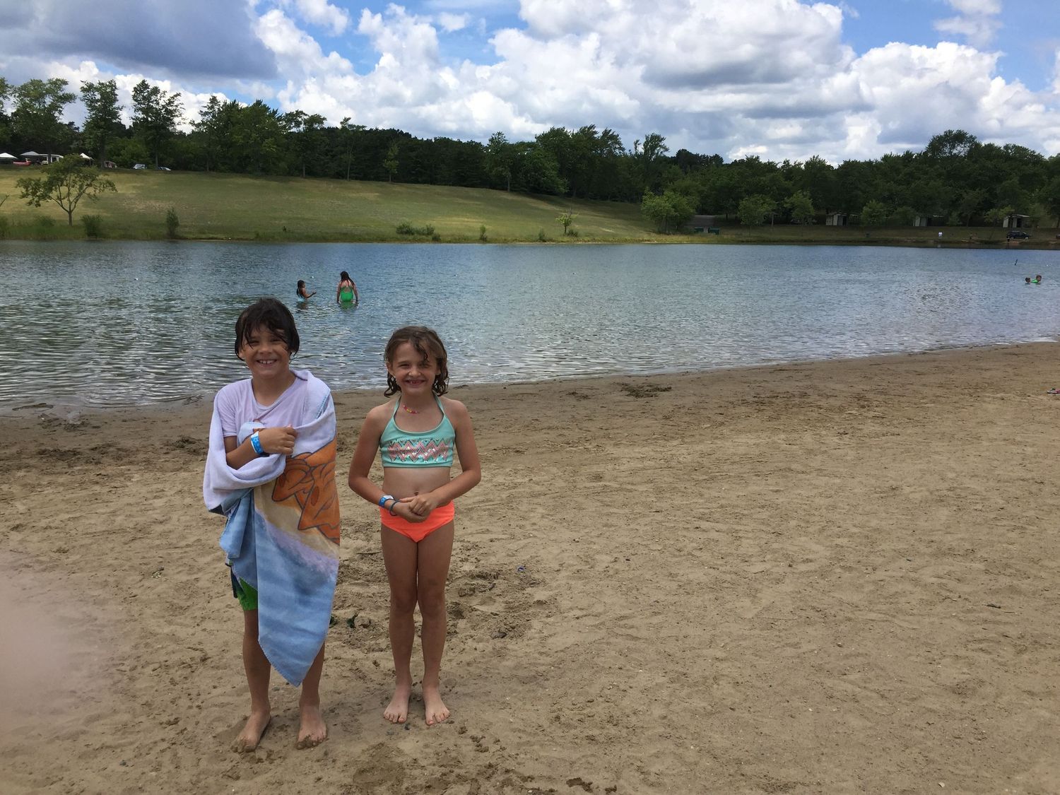 Two children stand smiling on a sandy beach by a calm lake on a cloudy summer day.