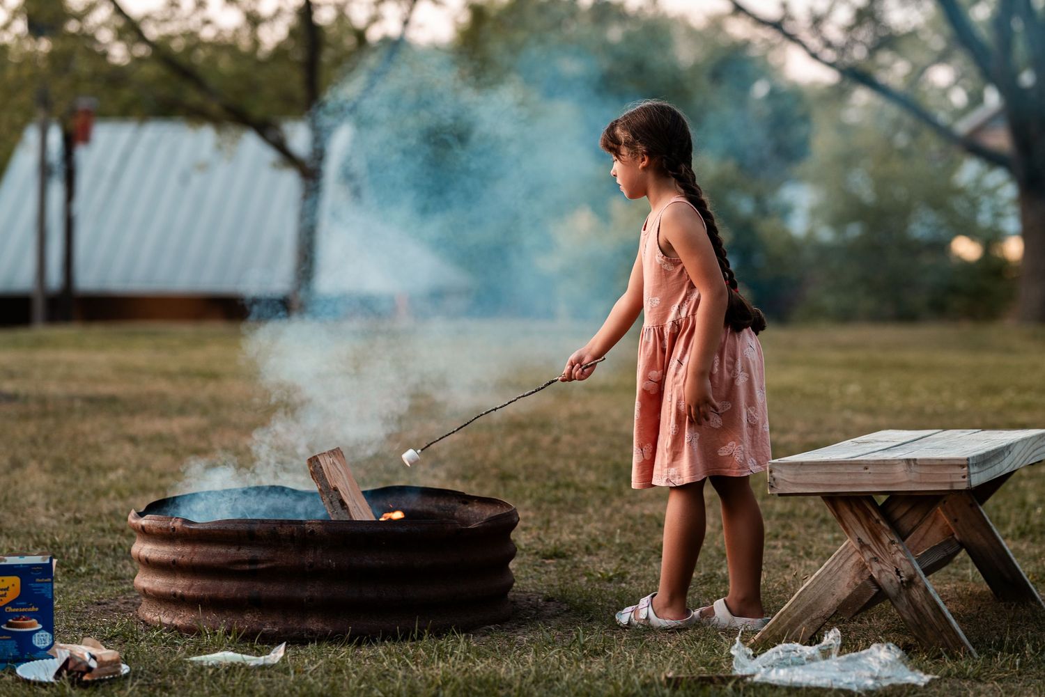 A young girl in a pink dress roasting marshmallows over a rustic fire pit outdoors at dusk.