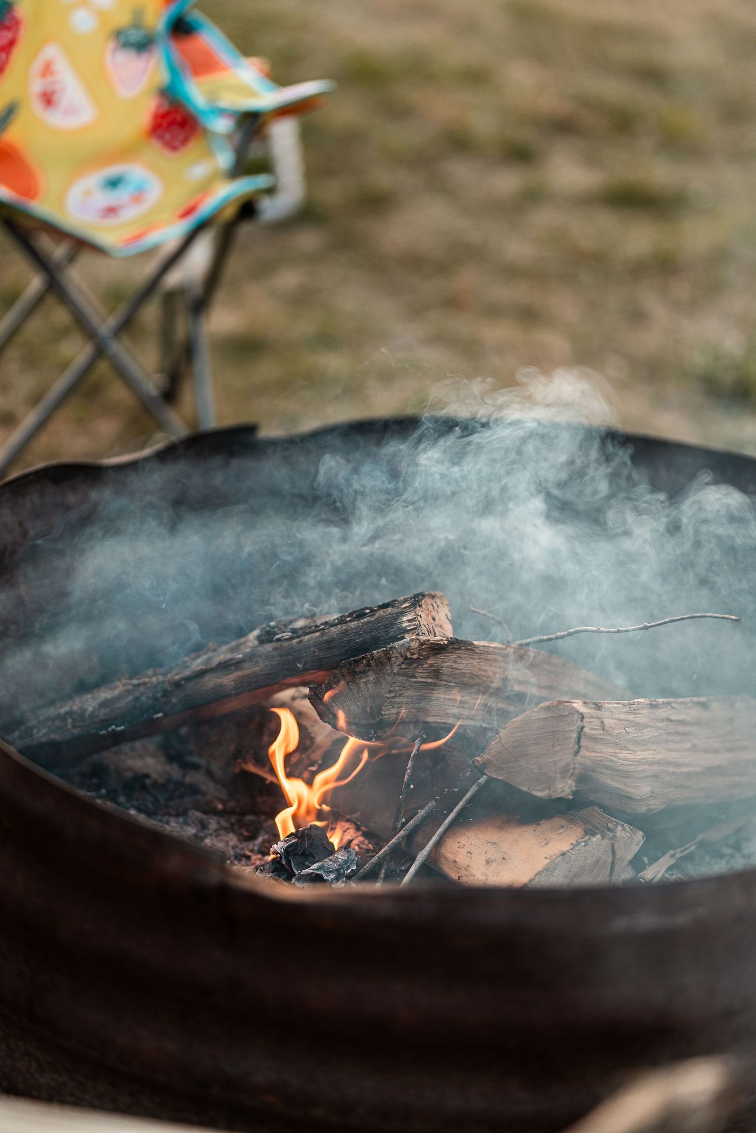 A crackling campfire burns in a metal fire pit with smoke rising, and a colorful folding camp chair visible in the background.