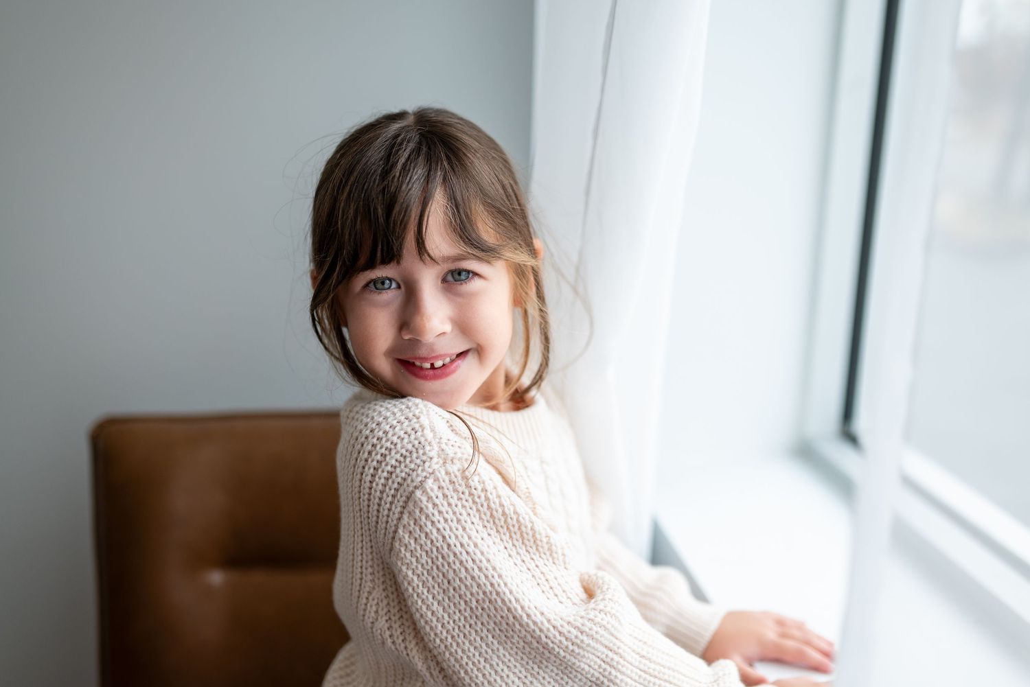 Someone in a cream knit sweater smiles while sitting by a bright window in a brown leather chair.