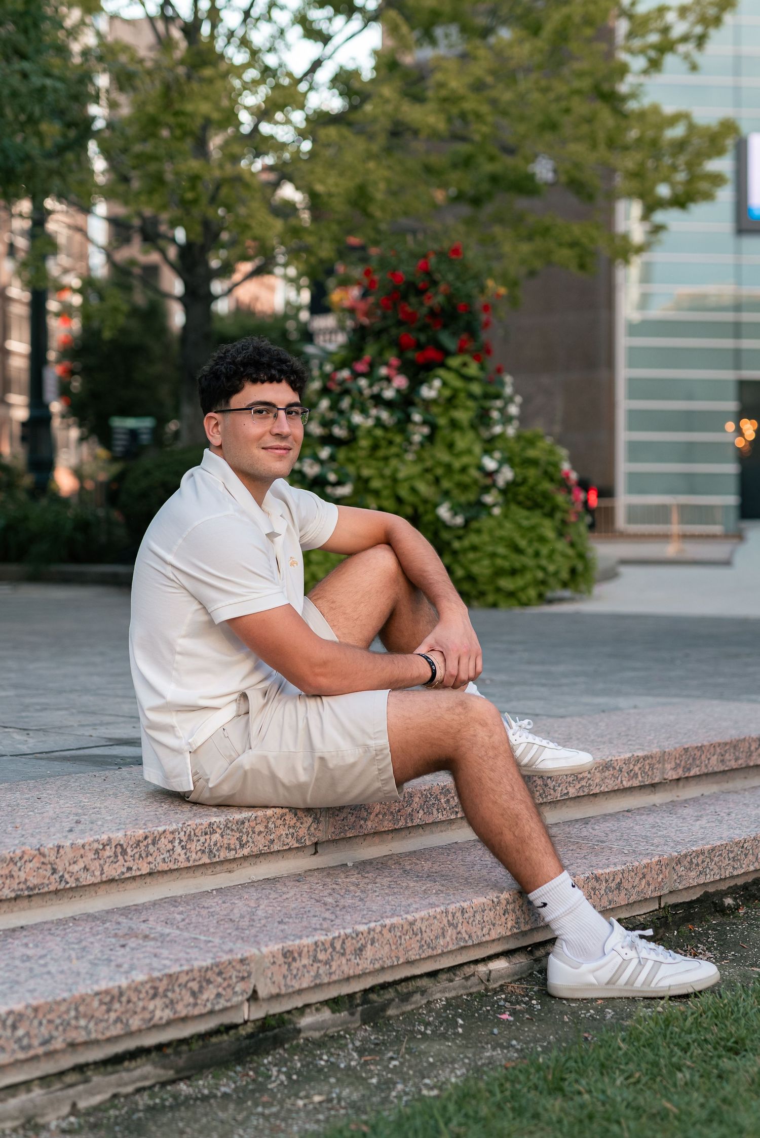Young man in white outfit sitting on stone steps outdoors, smiling, with lush green trees and red flowers behind him.