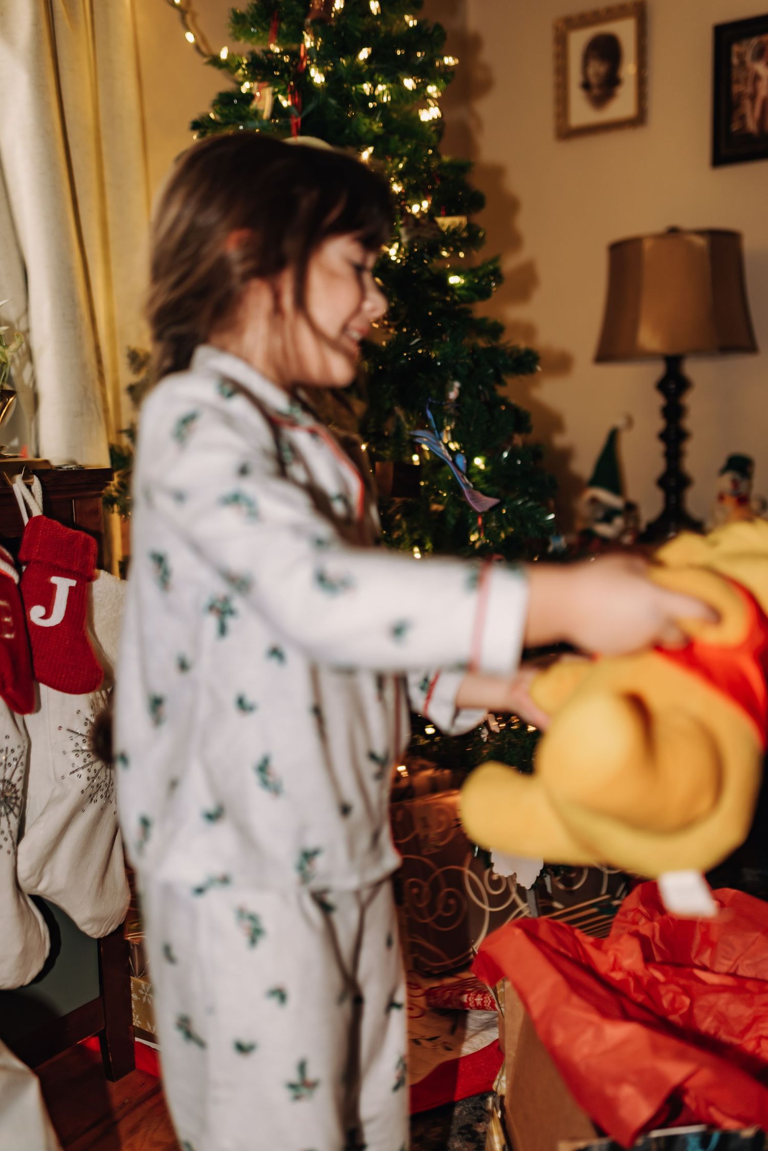 Child in festive pajamas reaching for decorations near an illuminated Christmas tree.
