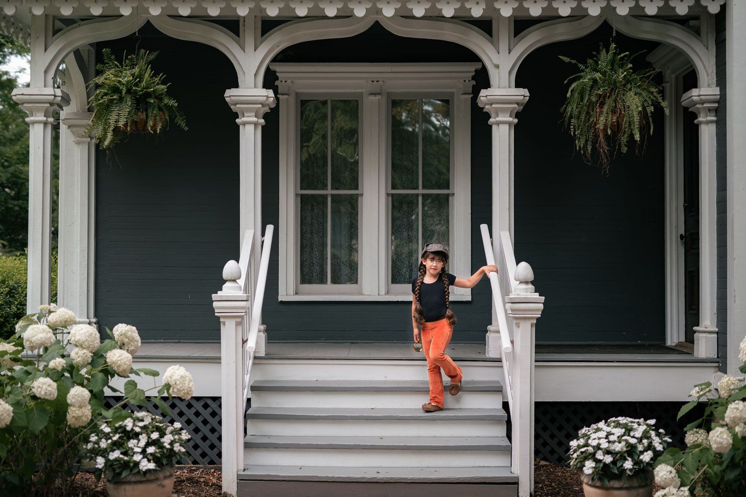 Child in orange pants poses on steps of charming Victorian porch with white columns and hydrangea blooms.