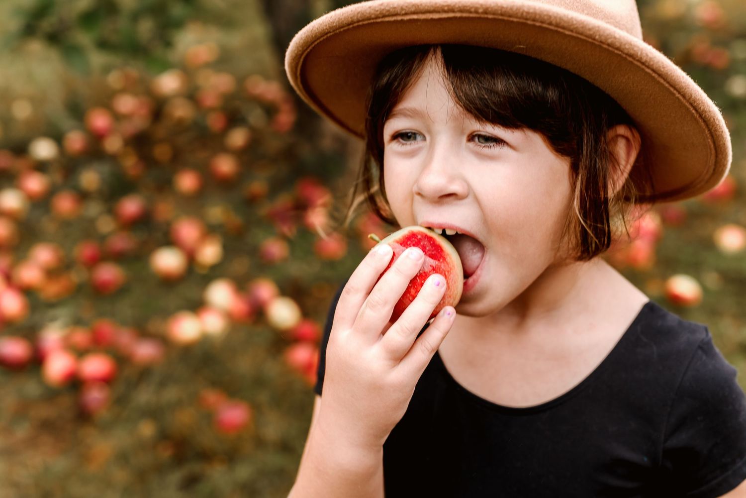 Someone wearing a brown hat takes a bite of a fresh red apple in an autumn orchard setting.