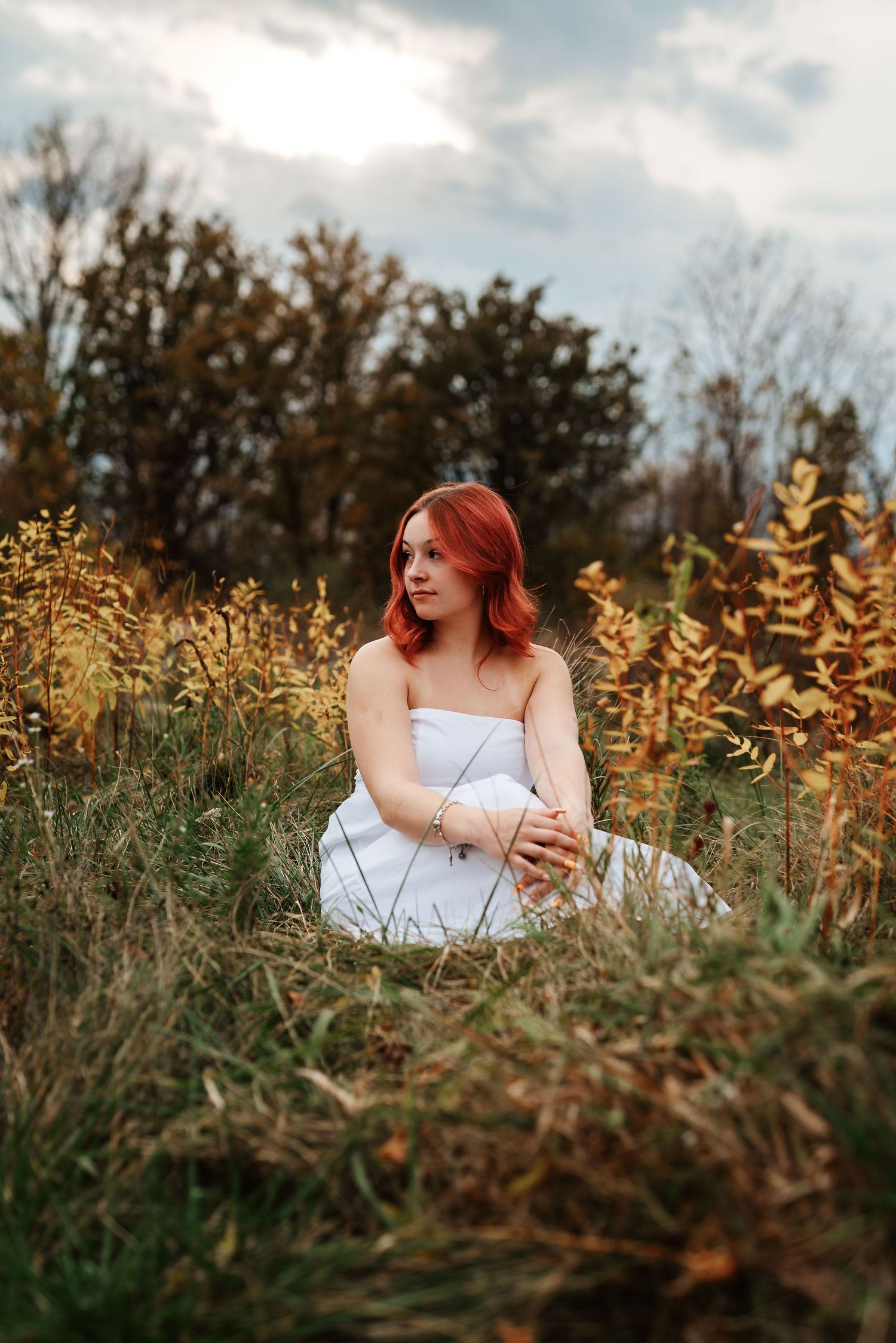 Young woman with vibrant red hair in white dress sitting among tall grass in scenic fall meadow.