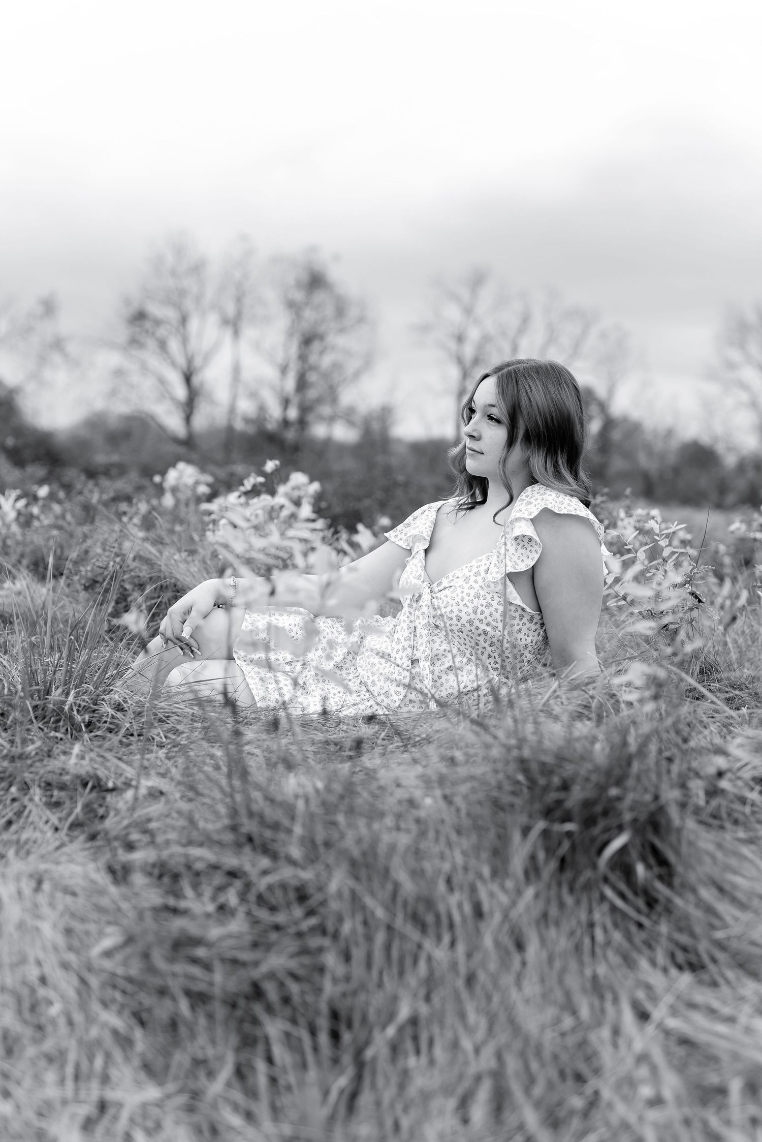 Young woman in floral dress sitting gracefully in tall grass field in black and white photo.