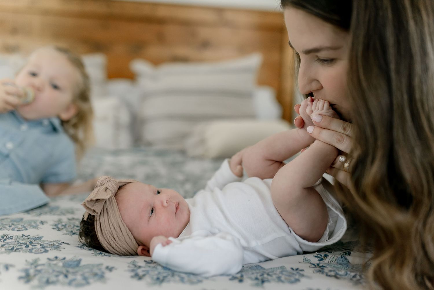 Mother kissing newborn baby's hand while toddler plays in background on bed.