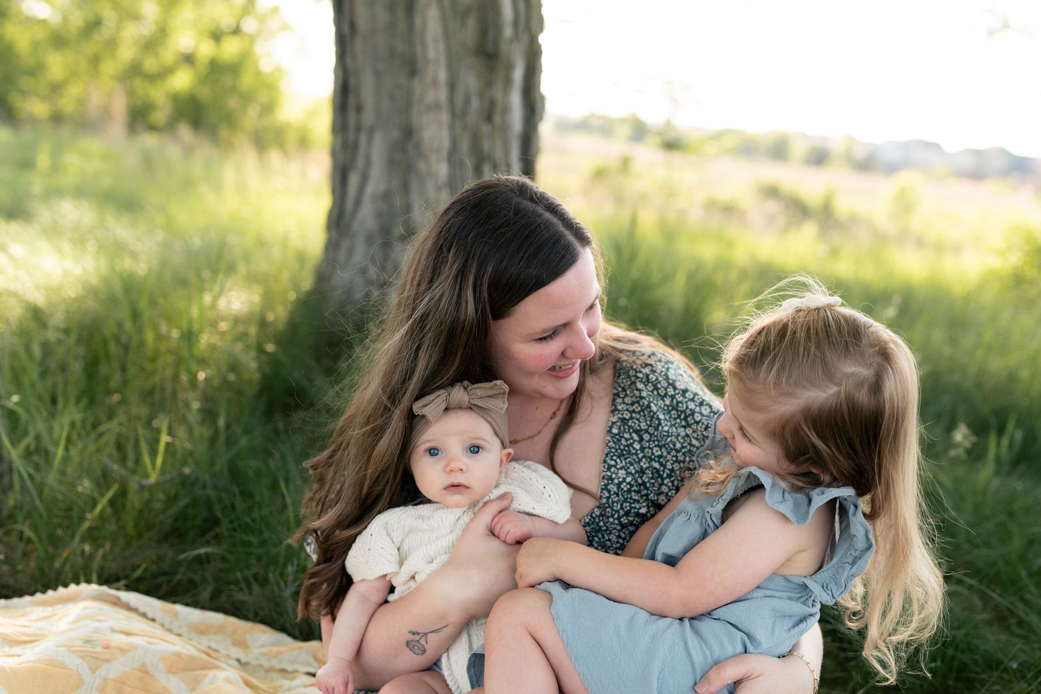 A mother sits outdoors on a blanket, smiling at her newborn baby while her young blonde daughter looks on.