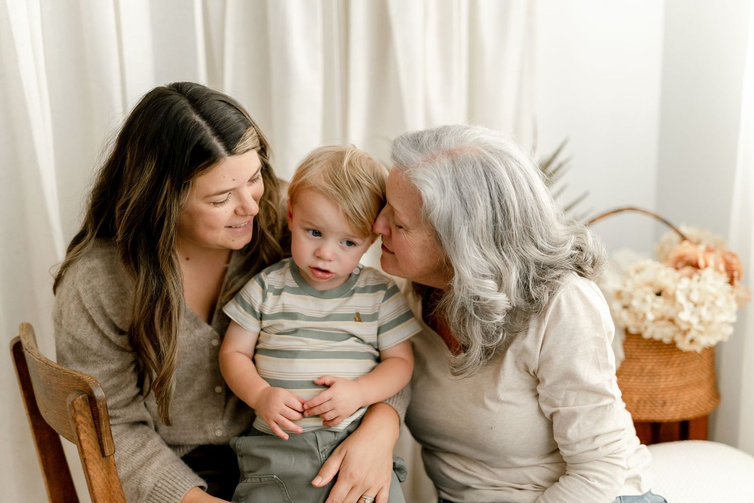 Two women with a toddler in a striped shirt sit close together in a bright room, smiling and sharing a tender family moment.