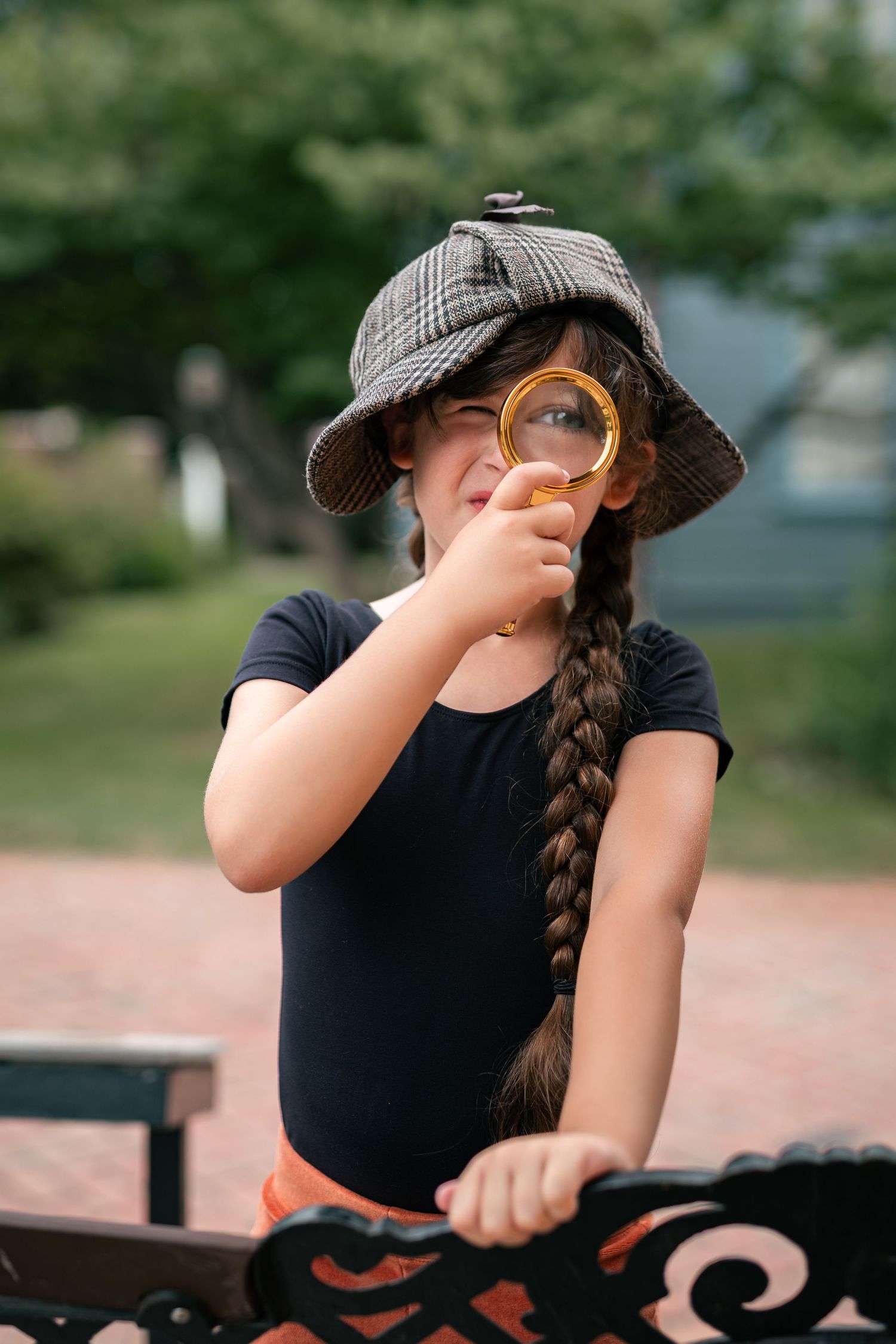 Young girl in detective hat holds magnifying glass to eye outdoors, playing inspector in a park setting.