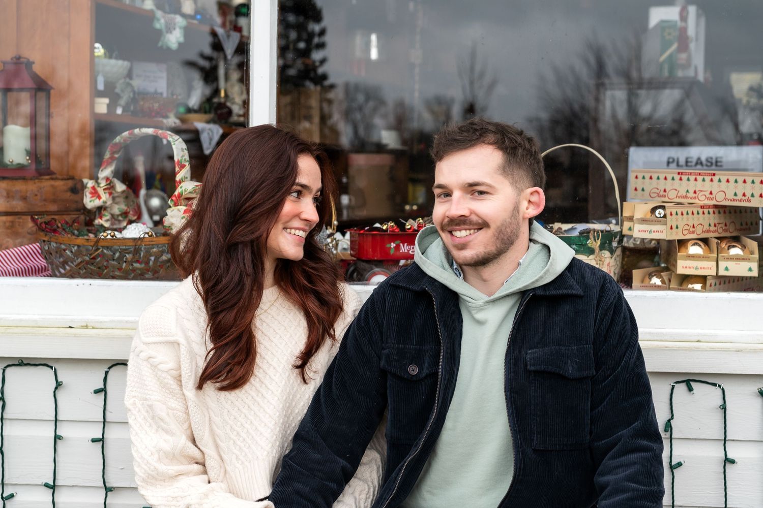 A couple shares a candid moment while sitting by a decorated storefront window during winter.