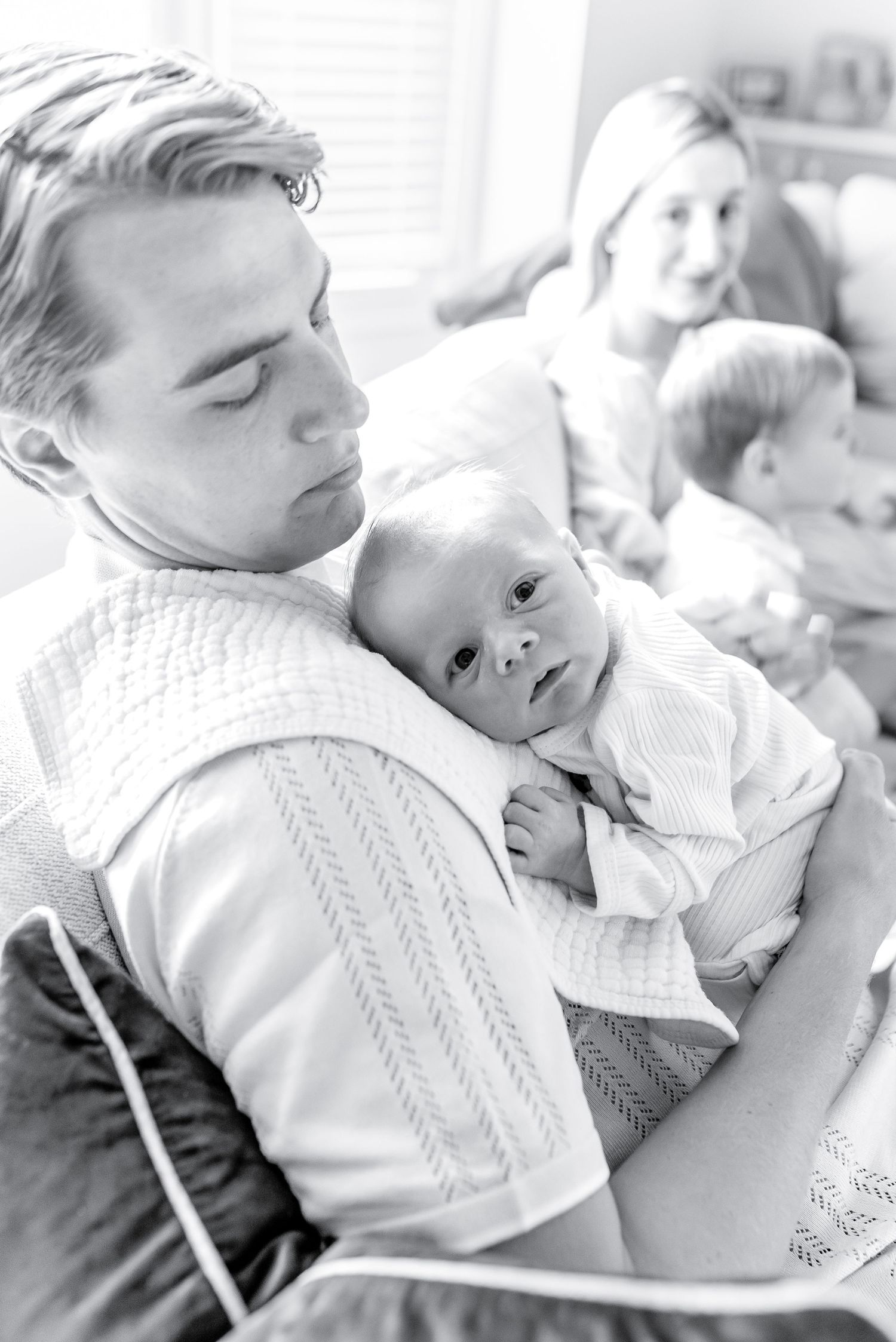 A tender black and white family moment shows a parent in a cable knit sweater holding a small baby while others look on.
