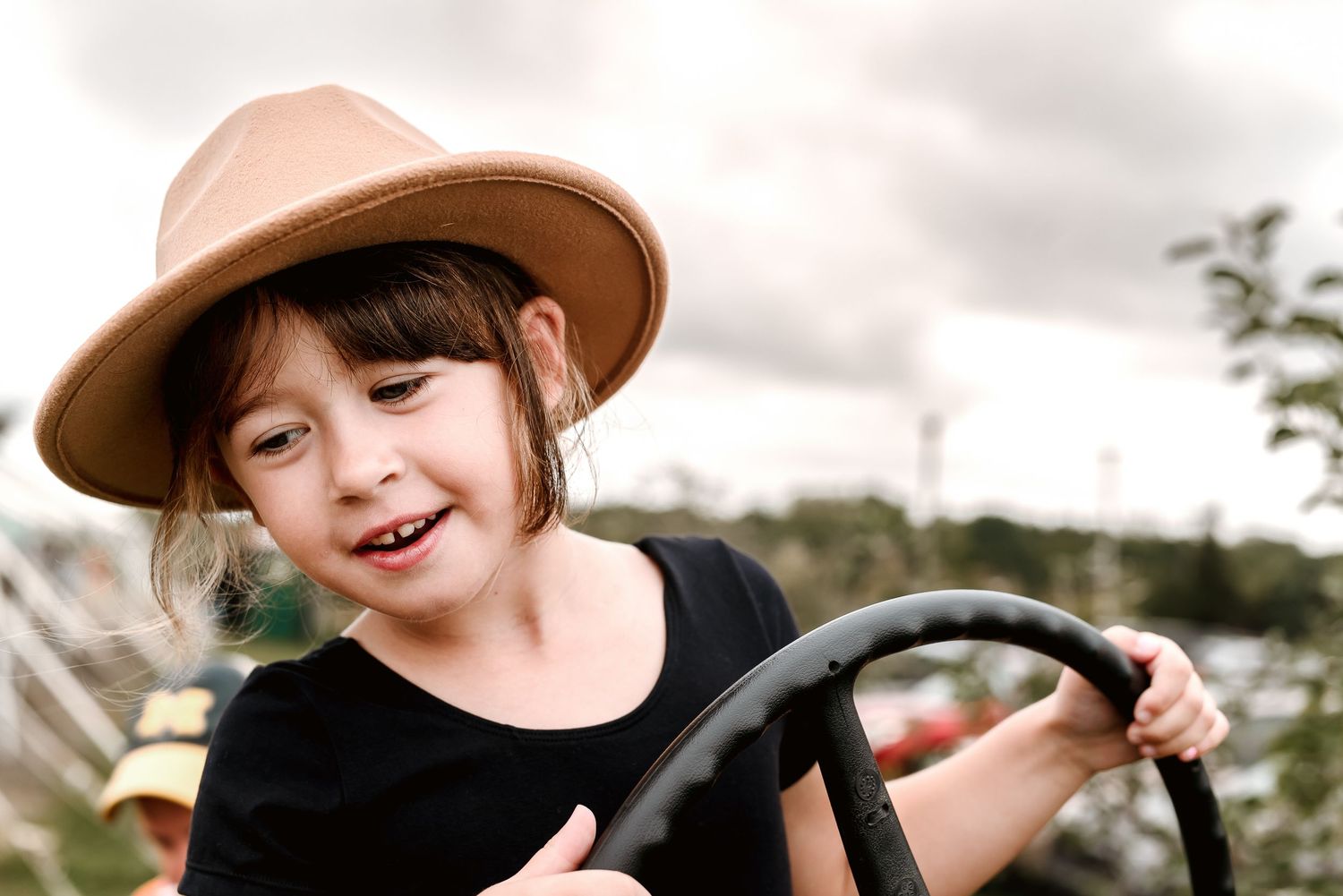 A child wearing a brown cowboy hat smiles while playing outdoors on a sunny day.