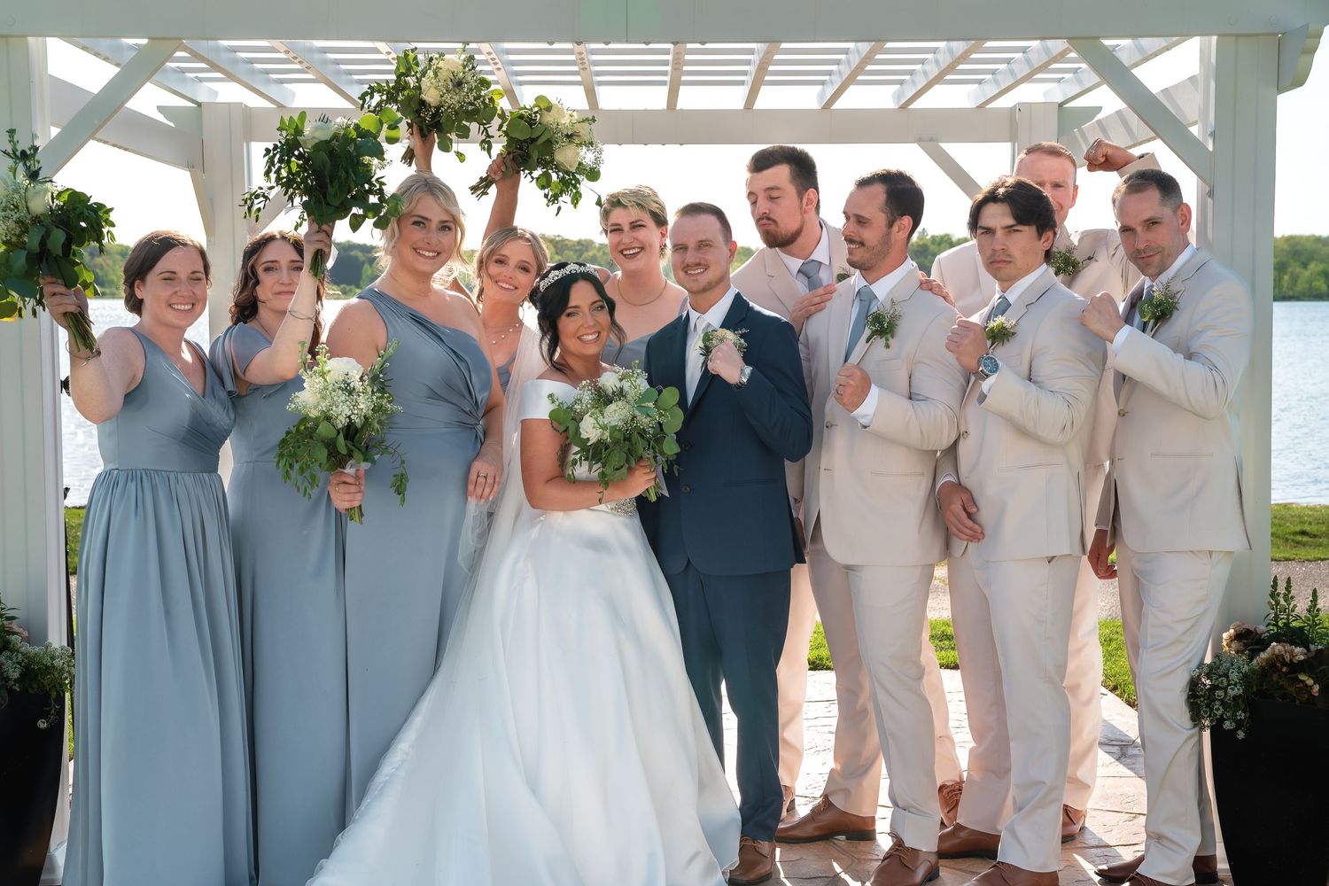 Wedding party poses under white pergola with bridesmaids in blue dresses and groomsmen in tan suits.