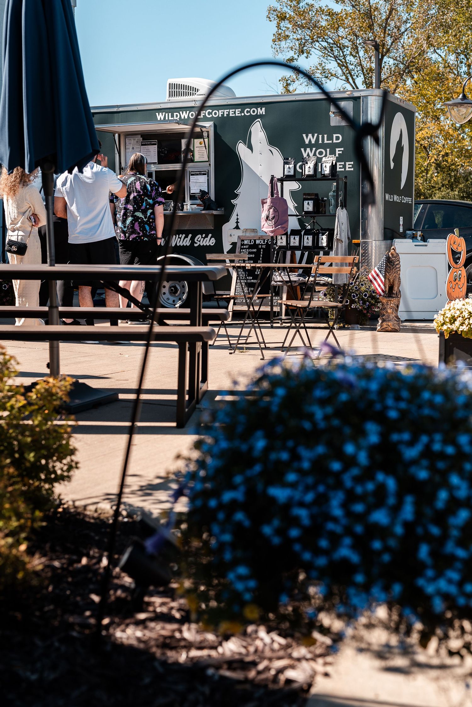 Wild Wolf Coffee food truck with customers ordering outdoors on a sunny day, surrounded by blue flowers and autumn decor.