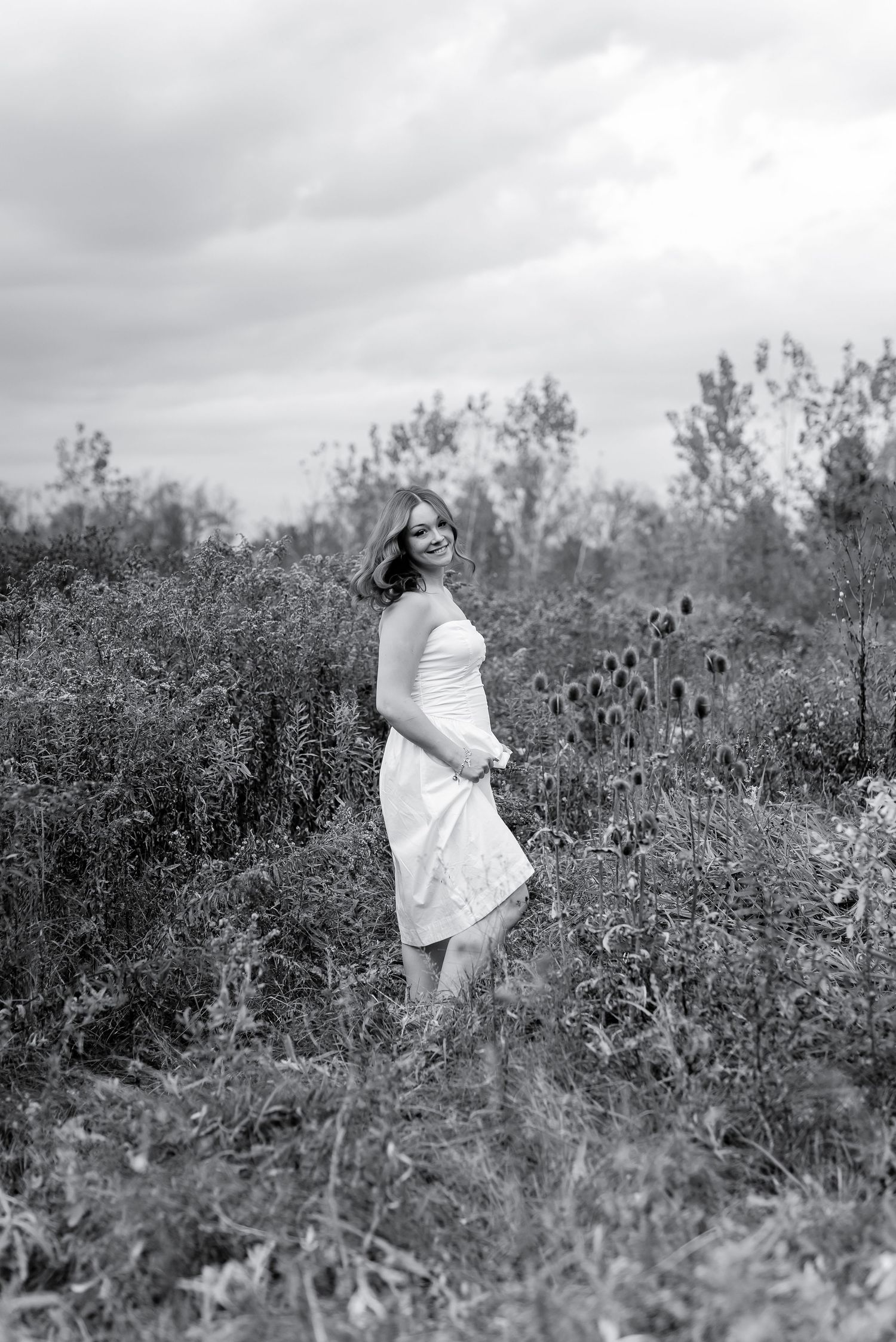 A woman in a white dress smiles while standing in a wild field, captured in black and white.