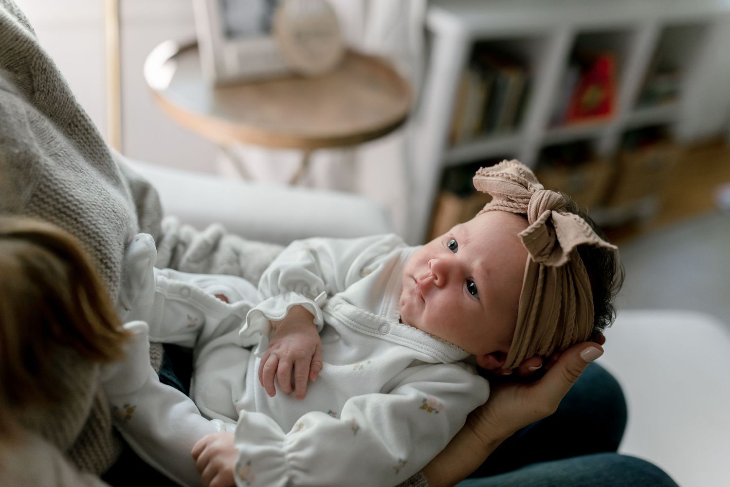 Newborn baby girl in white onesie with bow headband lying in mother's arms, gazing upward in cozy home setting.
