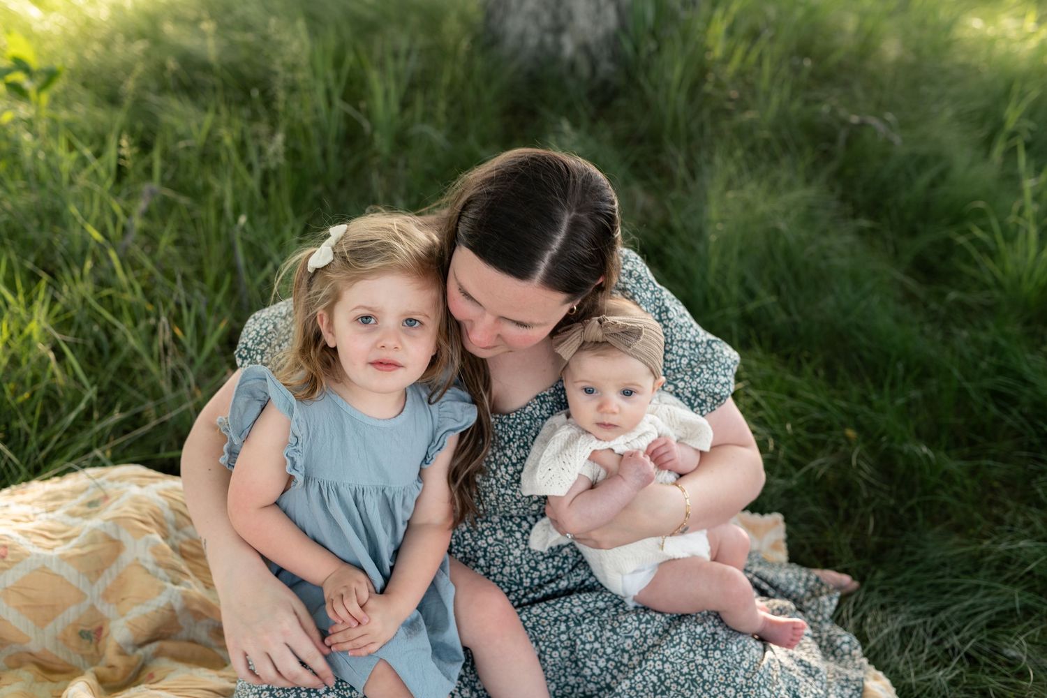 Mother sitting in green grass holding infant and toddler daughter in blue dress during outdoor family photo session.